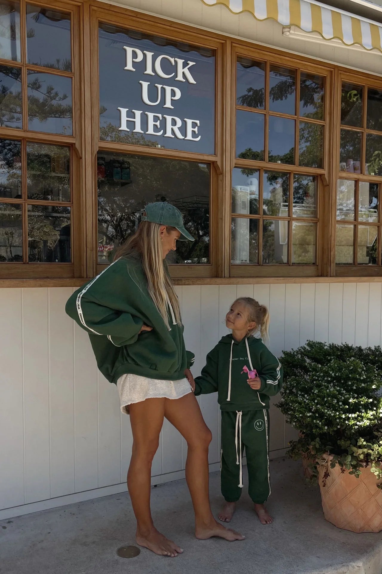 A woman and a young girl in matching green hoodies and CRACKER DAY CLUB Cracker Day Club - Kids Cloud Racer Sweatpants Forest stand barefoot outside a “Pick Up Here” sign, as the girl holds a pink toy and they smile at each other.