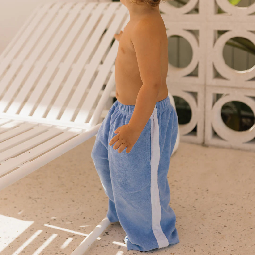 A toddler stands shirtless next to a white lounge chair, sporting GOLDEN CHILDREN's Ciao Tutti Relaxed Pants in Pool Blue. Circular designs on the wall enhance the bright outdoor patio ambiance.