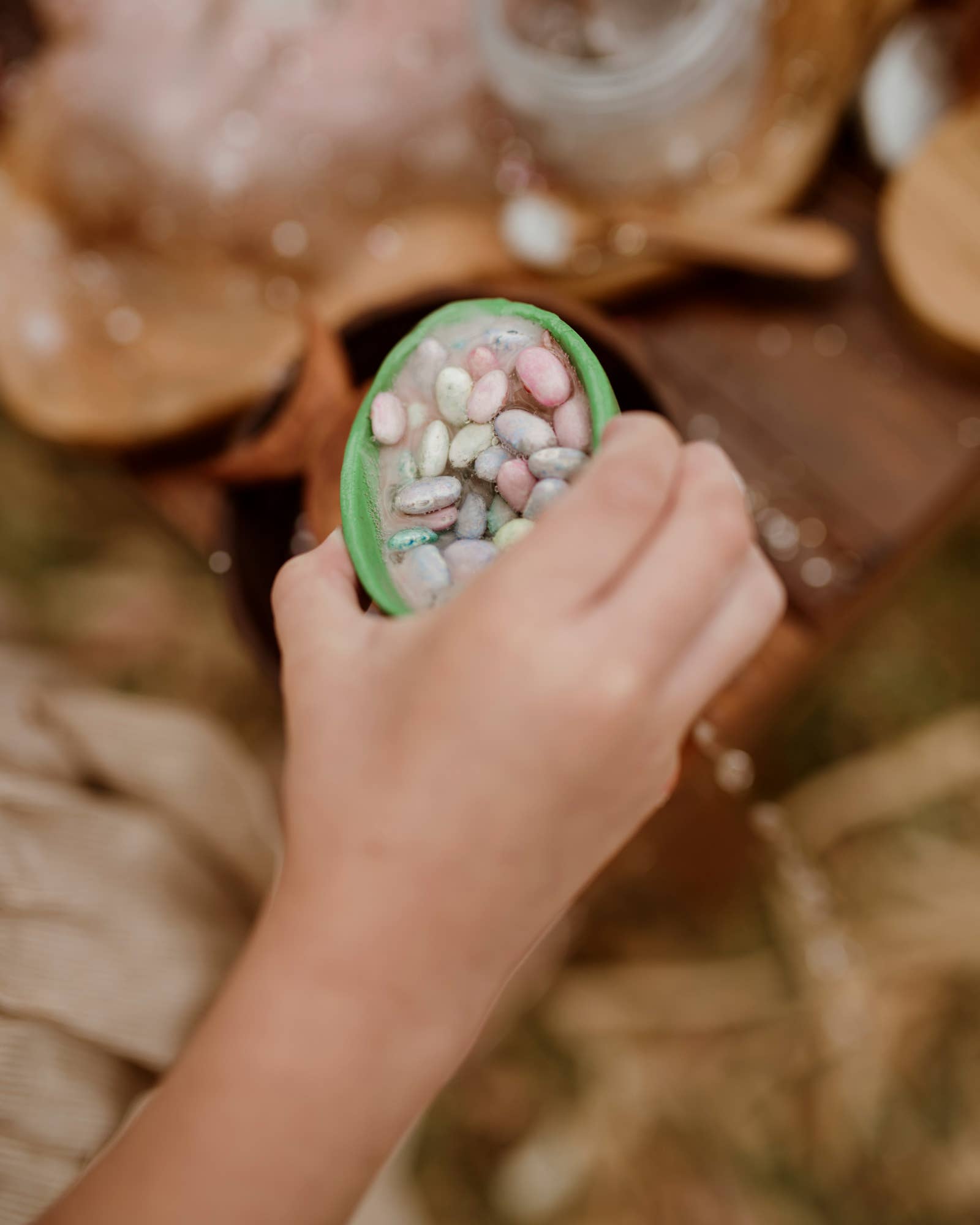 A hand holds a green cup filled with colorful small stones from The Little Potion Co's MINI Easter Egg Hunt Potion Kit, by THE LITTLE POTION CO. A blurred background shows a wooden surface and some leaves, creating an enchanting outdoor ambiance.