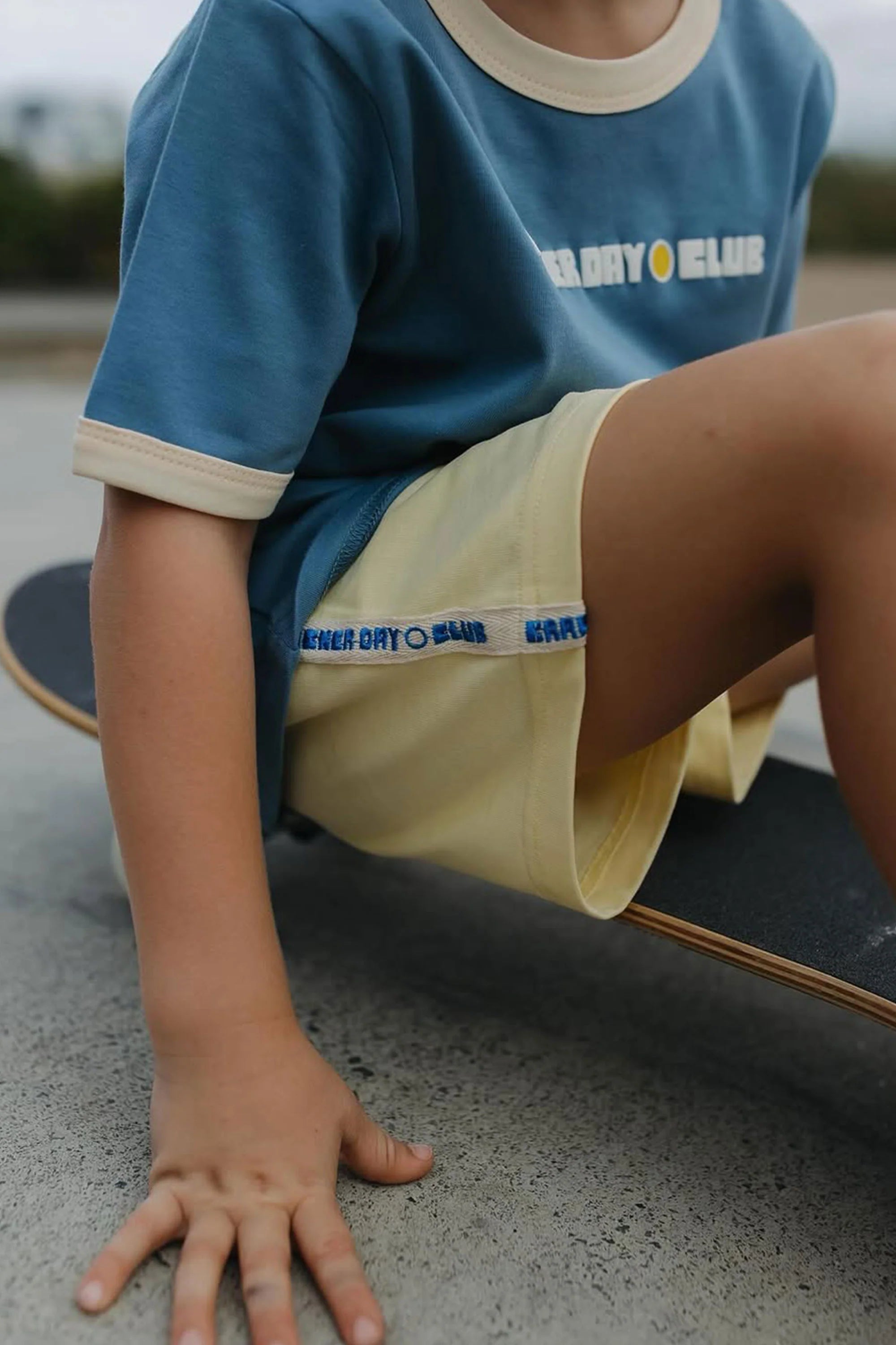 A young child wearing CRACKER DAY CLUB's Cracker Day Club - Kids Racer Club Shorts Sunshine and a blue "EARLY DAY CLUB" tee sits on a skateboard outdoors. The cotton shorts feature retro stripes and are perfect for active play.