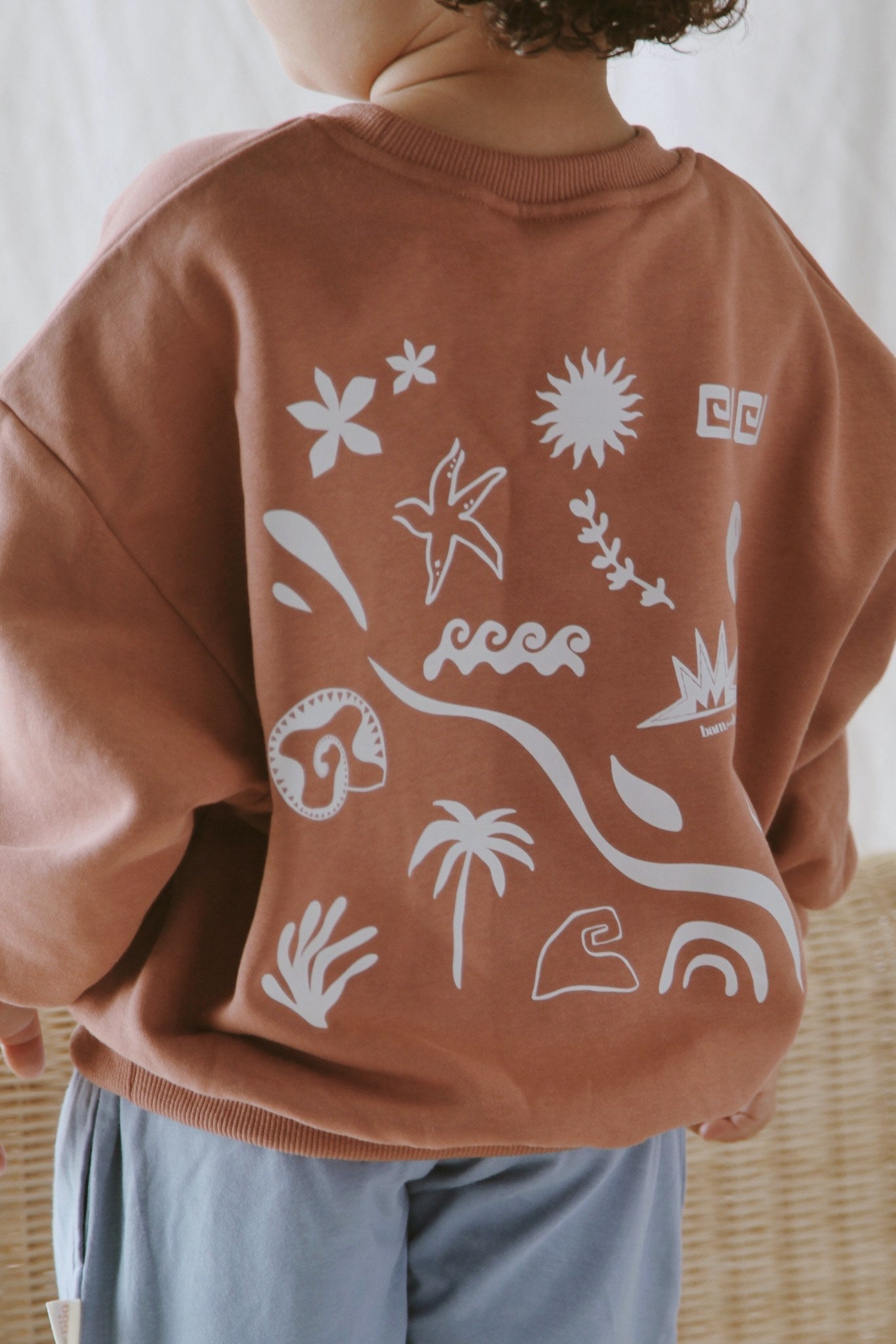 A child wears the BAM LOVES BOO Beachcomber Jumper Clay—a brown organic cotton, unisex boxy-fit jumper with white abstract nature designs (palm tree, sun, waves, bird, and rainbow)—seen from the back near a wicker chair.