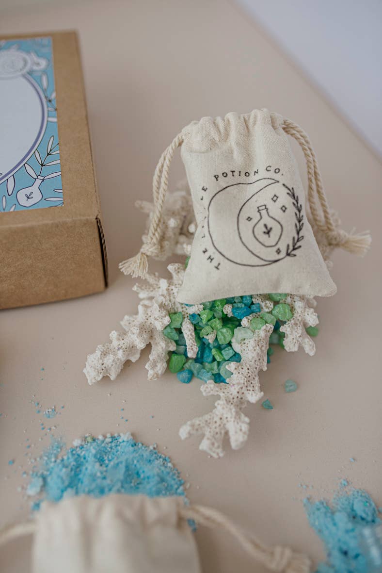 A small burlap pouch with "THE LITTLE POTION CO" logo lies open, spilling colorful green, blue, and white crystals onto a light surface. Nearby, there's more blue powder and a cardboard box labeled "Moonlight Waters MINDFUL Potion Kit." White coral pieces are scattered among the crystals in this mystical potion kit.