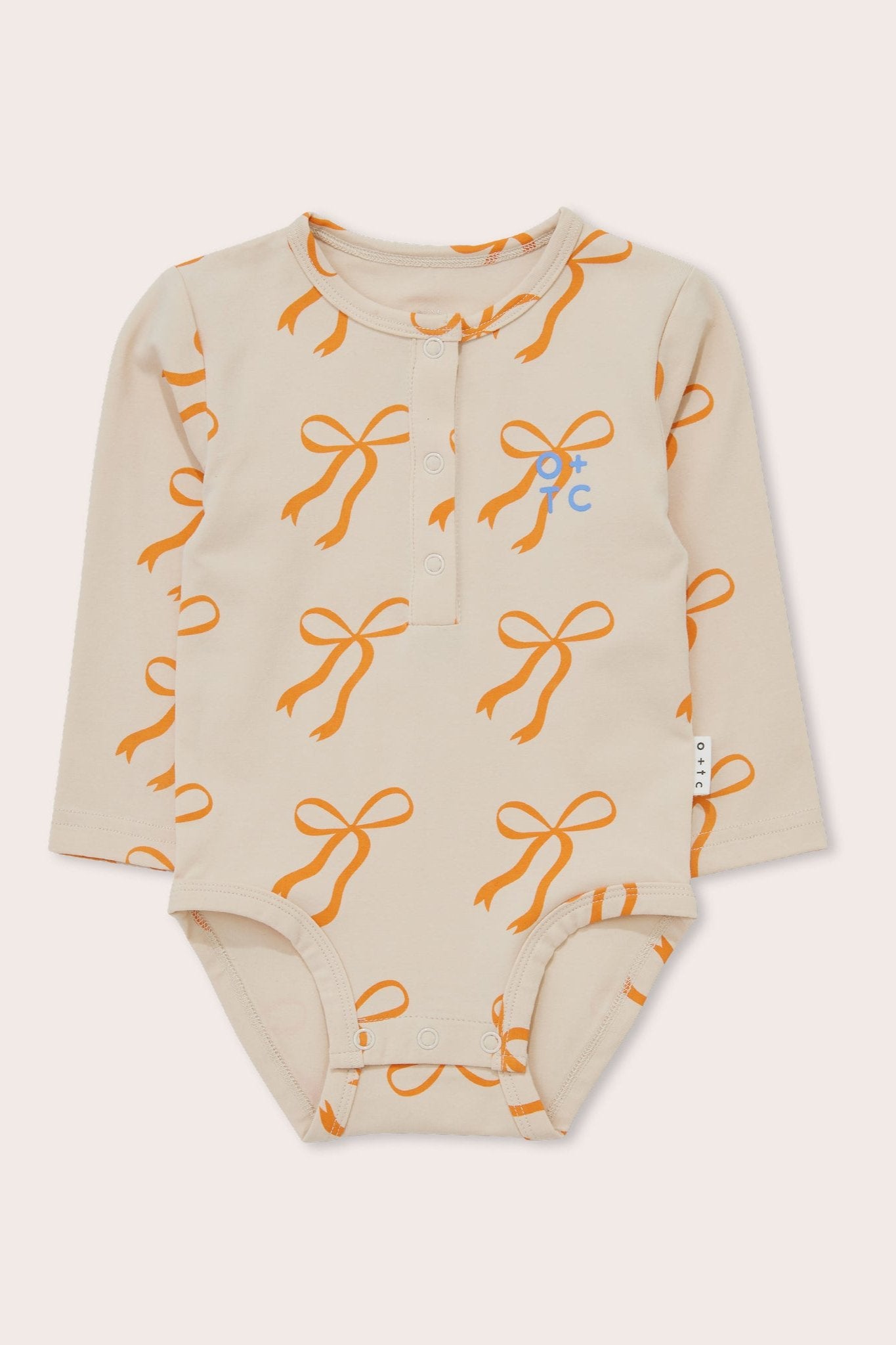 A cream-colored long-sleeve bodysuit for babies, featuring a front button closure and accented with golden bows. The Cream Bows Long Sleeve Bodysuit from OLIVE + THE CAPTAIN is made from stretch cotton terry and includes a small blue logo on the left chest along with a white brand tag on the right side seam.