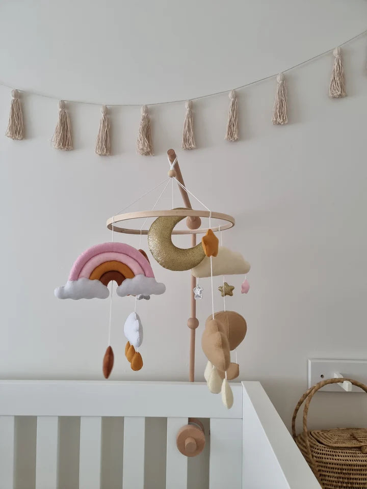 The ROOMI & ME Roomi & Me - Baby Mobile Sparkly Galaxy features plush rainbow, moon, stars, clouds, and raindrops for gentle visual stimulation above a white crib, with a beige tassel garland on the wall behind.