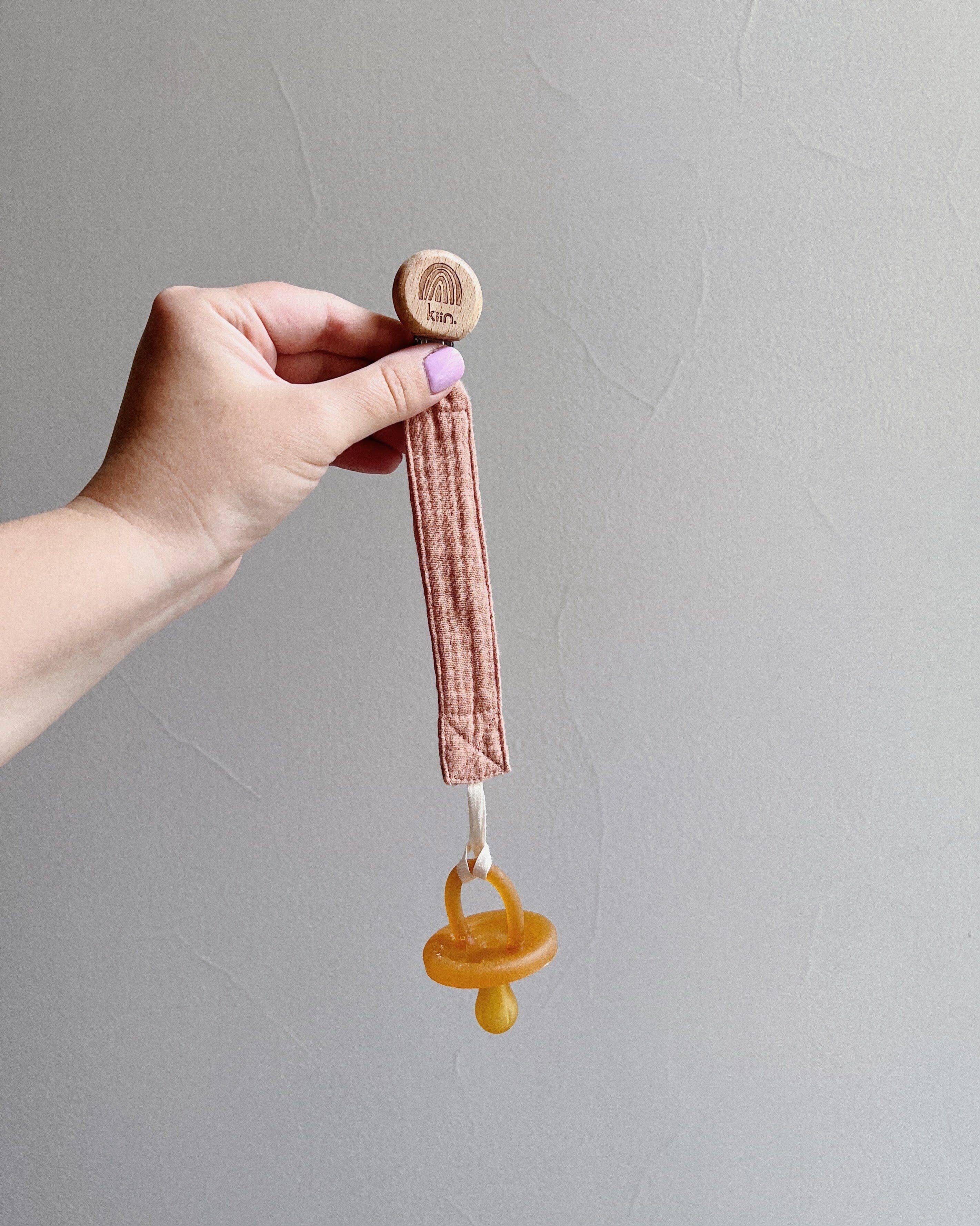 A pacifier clip with a wooden clasp reading "Kiin Baby" is pictured, featuring a mustard silicone pacifier. Crafted from pink fabric, the Cotton Dummy Clip Oat makes an ideal gender-neutral baby shower gift, shown against a plain, light-colored wall.