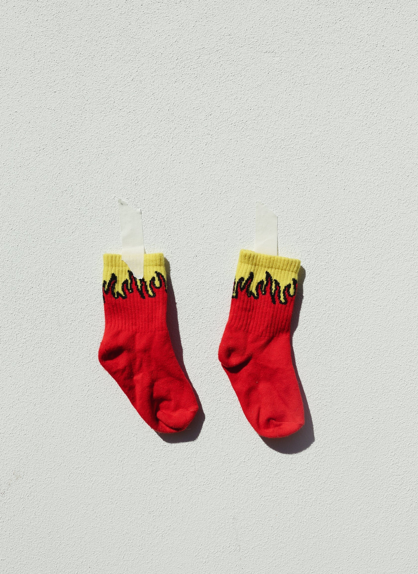 A pair of TINY LOVE CLUB's Fire Socks, showcasing a sports-style design with yellow and black flame patterns at the top, are taped to a light gray wall. The red socks, which feature elastic-banded ankles, are positioned side by side, enhancing the clean, minimalistic look of the image.