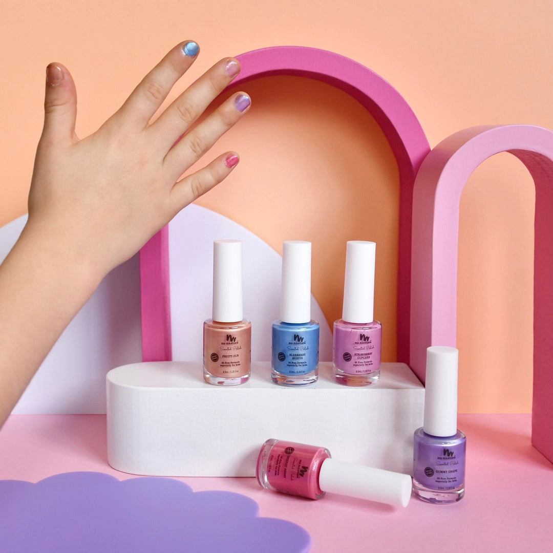 A hand with pastel nails is shown above four bottles of NO NASTIES Scented Scratch Off Kids Nail Polish Strawberry Cupcake in Pastel Pink, set on a white display with pink and purple arches in the background.