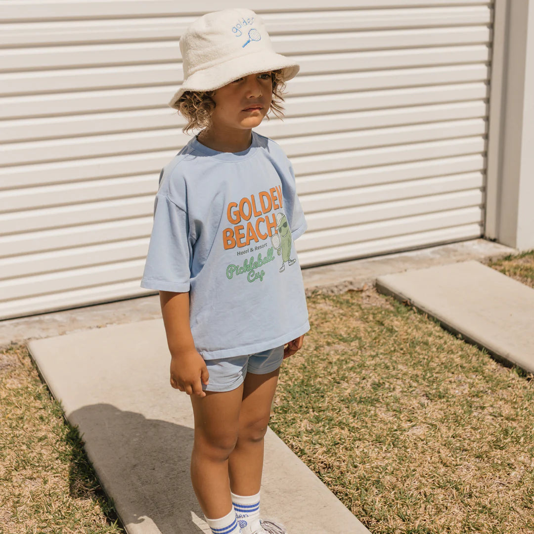 A young child stands outdoors in a comfy cotton jersey "Golden Beach" t-shirt by GOLDEN CHILDREN, with Retro Boy Shorts Palm Blue and a white bucket hat. The relaxed fit makes play easy on the pathway by grass, with a closed white garage door in the background.