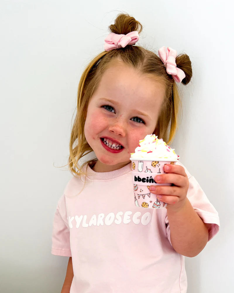 A young girl with two pink bows smiles while holding the BBcino - Reusable Babycino Cup 120ml Tough Cookie in Pink by BBCINO. She wears a light pink "KYLAROSECO" shirt against a plain white background. The cup is topped with whipped cream and sprinkles.