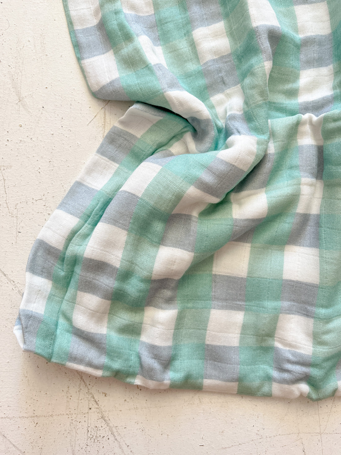 The Milky Designs ~ Seamist Gingham Wrap by MILKY DESIGNS, featuring a white, light blue, and mint green checkered pattern, lies crumpled on a textured white surface, highlighting its exceptional breathability.