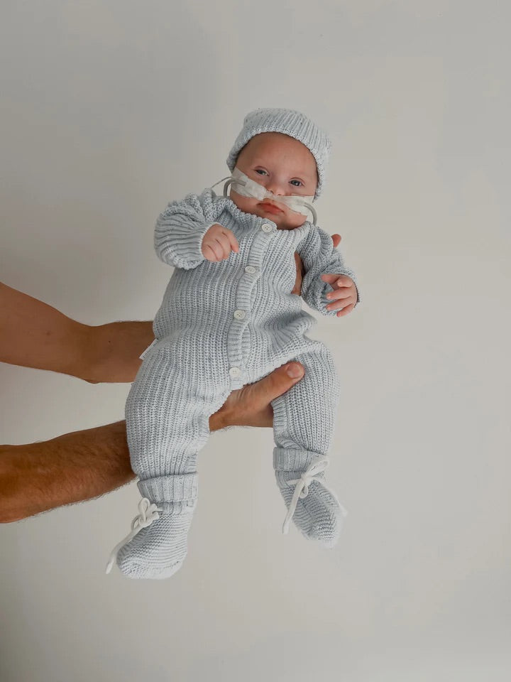 A baby with a nasal feeding tube is held up by an adult, wearing the ZIGGY LOU Classic Knit Romper in Aero Fleck, against a plain light background and looking at the camera.