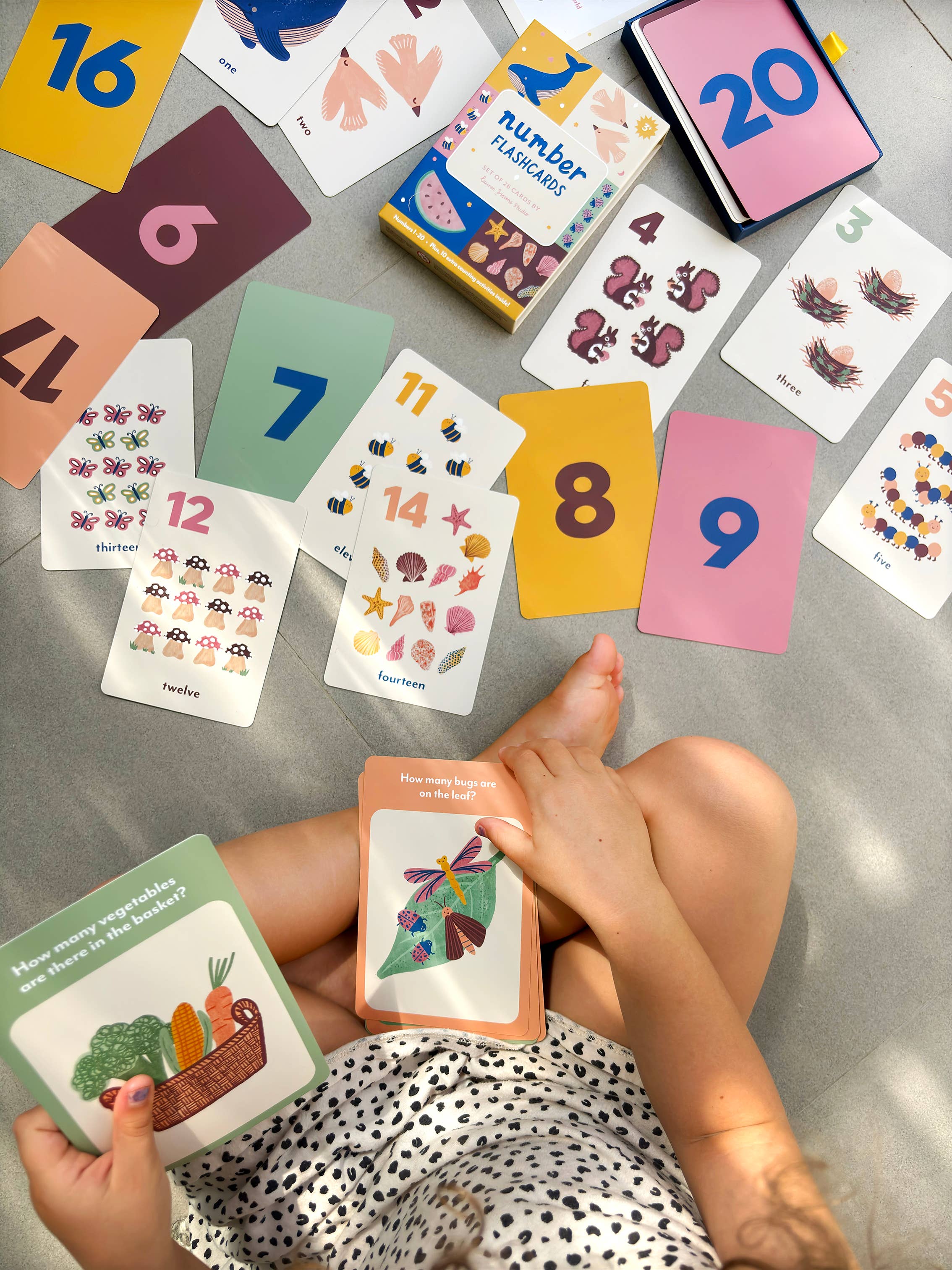 A child sits on the floor, playing with Lauren Sissons - Illustrated Number Flashcards by Lauren Sissons Studio, holding cards featuring vegetables in a basket and a bird, building counting skills through playful learning.