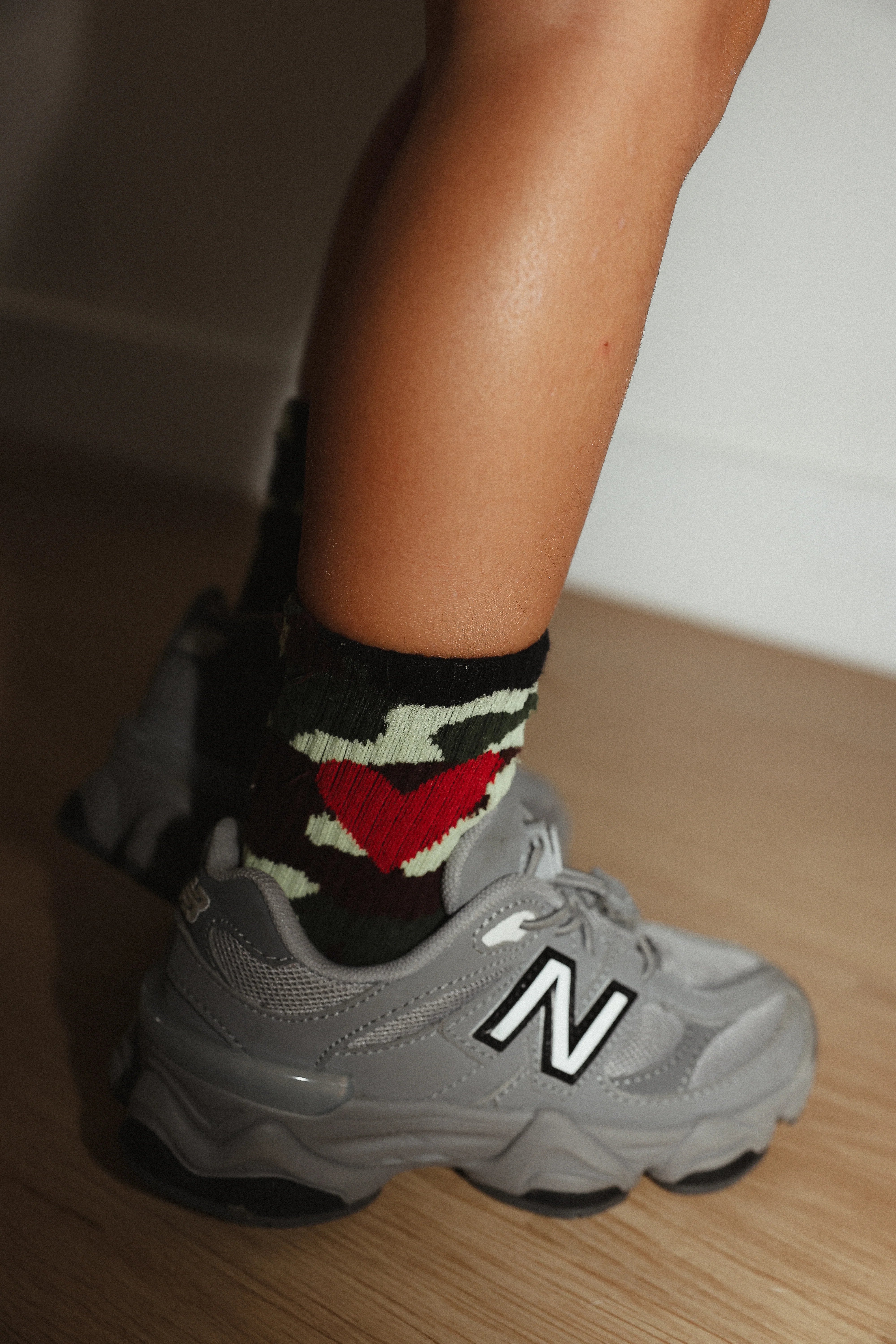 A person wearing gray New Balance sneakers and TINY LOVE CLUB's Camo Heart Socks, featuring a red heart, green camouflage pattern, and elastic banded ankles, stands on a wooden floor.