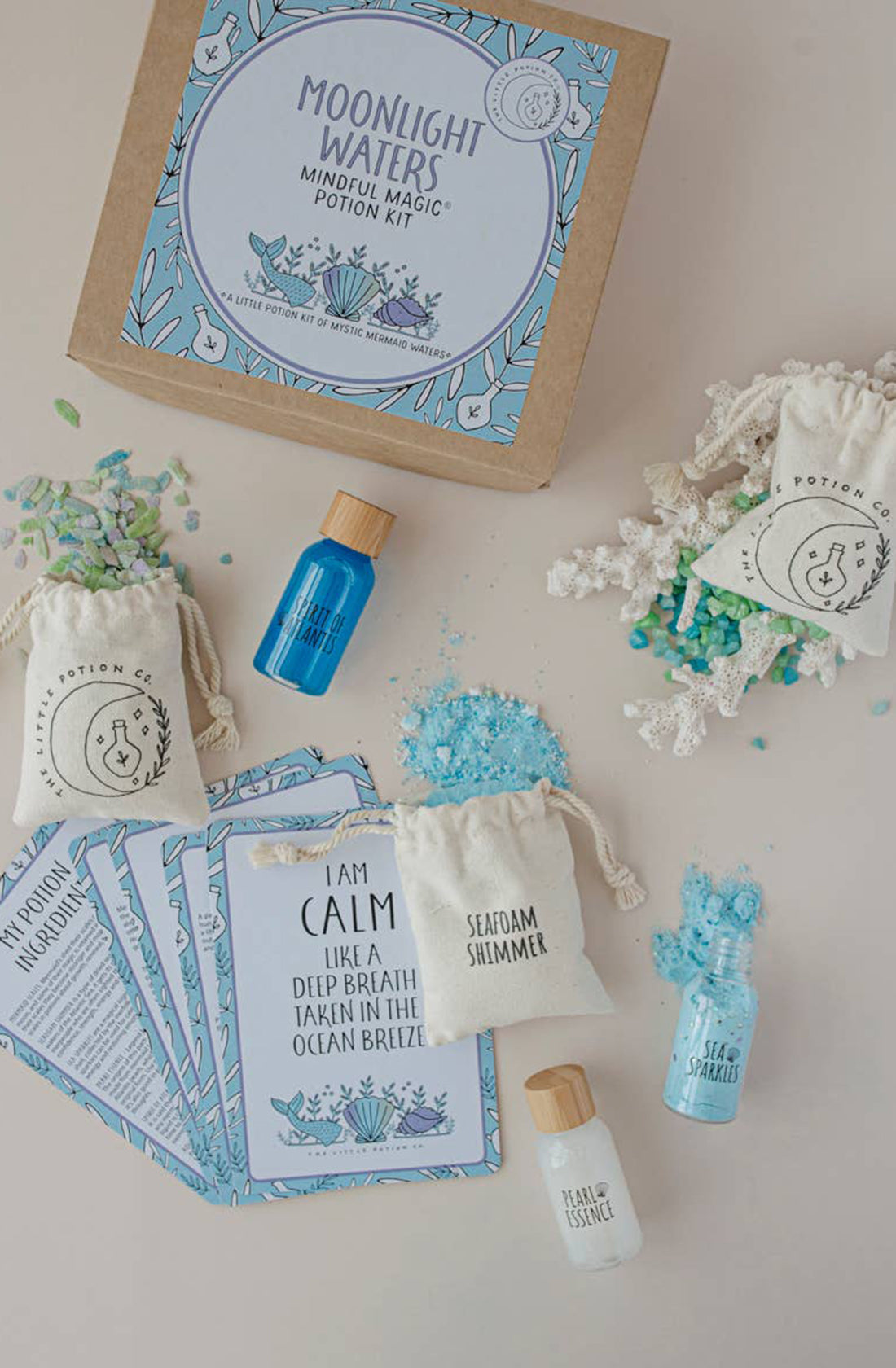 A "Moonlight Waters MINDFUL Potion Kit" from THE LITTLE POTION CO is displayed. This enchanting kit includes small bottles of colored liquids, sachets filled with blue crystals, and several cards featuring calming phrases. The items are neatly arranged on a light surface, creating a serene and organized layout perfect for sensory play.