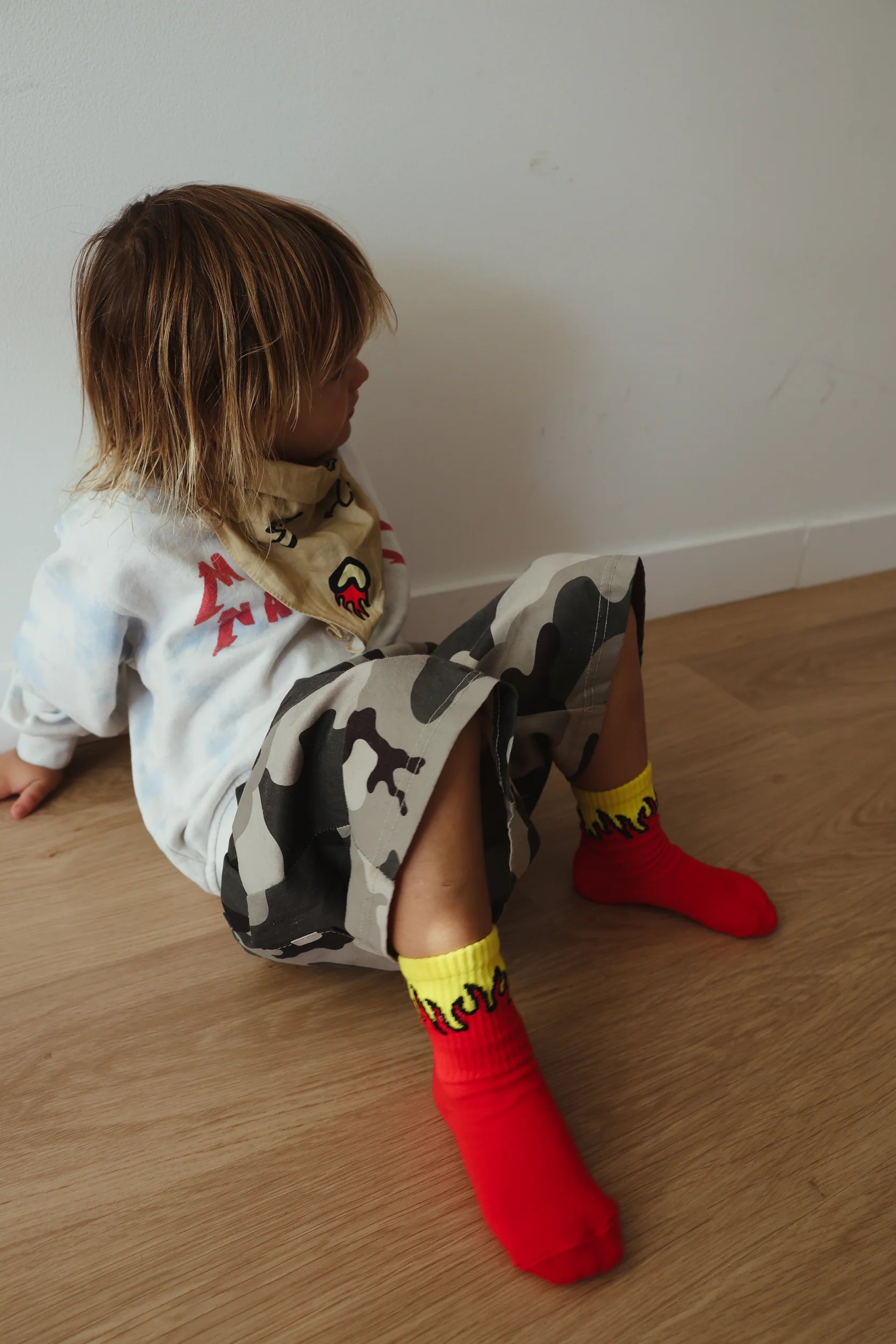 A child with blonde hair sits on the floor facing sideways, wearing a white shirt with red designs, camouflage shorts, and bright red TINY LOVE CLUB Fire Socks paired with elastic-banded ankles. A light-colored bandana featuring a cartoon face is tied around their neck. The background is a plain wall and wooden floor.