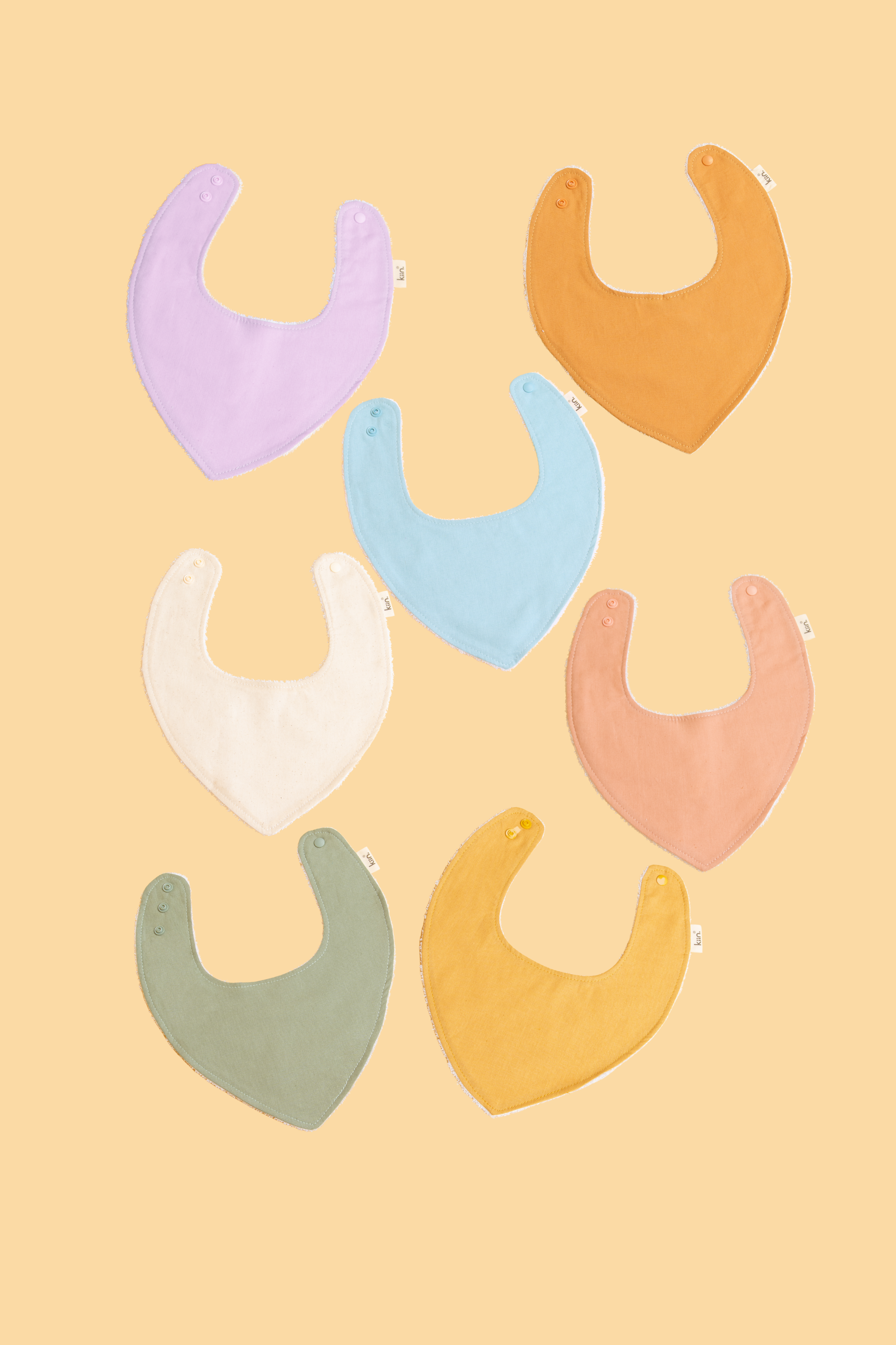 The KIIN BABY Dribble Bib Oat collection includes seven bamboo/cotton towelling bibs in purple, orange, blue, white, pink, green, and mustard yellow. They feature snap fasteners and are displayed in two rows against a beige background.