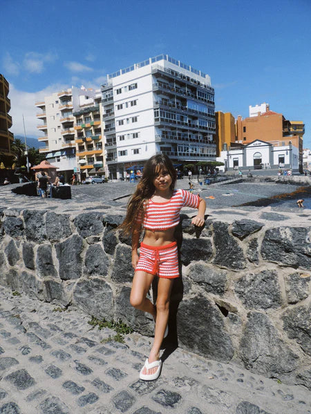 A young girl with long hair stands against a stone wall, wearing the Crochet Stripes Set Red/White from NOTHING BUT AMOR. Behind her are multi-story buildings and a clear blue sky, capturing the laid-back vibes of Bali on a bright, sunny day.