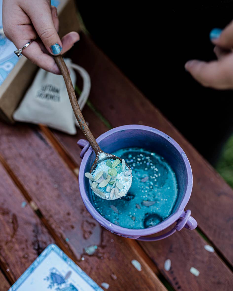 A person's hand, with blue-painted fingernails, holding a wooden spoon stirs a bluish liquid with white and green particles in a purple cauldron. The scene, evoking the mystical charm of the Moonlight Waters MINDFUL Potion Kit by THE LITTLE POTION CO, unfolds on a wooden surface with a partially visible card and a small beige pouch nearby.
