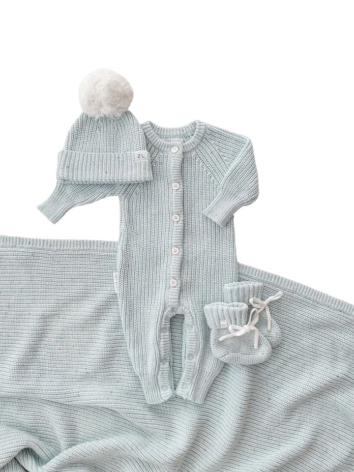 The ZIGGY LOU Classic Knit Romper in Aero Fleck features white buttons, a matching knit hat with a large white pom-pom, booties, and a coordinating Ziggy Lou knit blanket—all displayed on a white background.