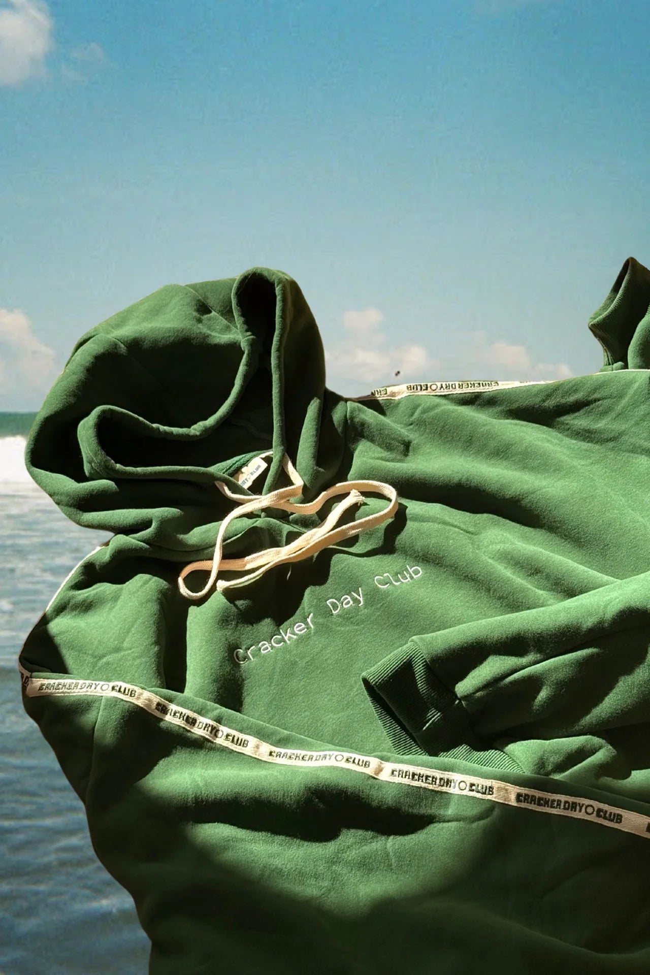 The CRACKER DAY CLUB Cloud Racer Hoodie Forest, made from heavyweight terry cotton for an oversized fit, features white drawstrings and "Cracker Day Club" text. It is displayed against ocean waves and a blue sky with clouds.