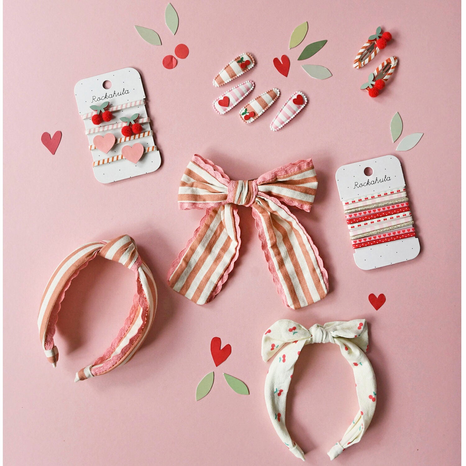 A flat lay of ROCKAHULA hair accessories, featuring the Rockahula - Stripy Cherry Ponies, striped bows, and clips in pink, white, and red, displayed on a pink background with heart and leaf cut-out decorations.