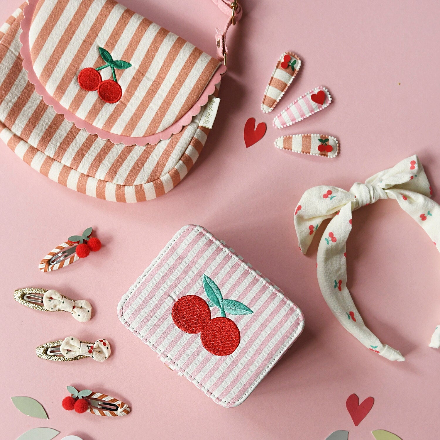 A pink background displays ROCKAHULA's Rockahula - Sweet Cherry Clips with grosgrain ribbon, a cherry-embroidered striped purse and box, a fabric headband with cherries, and heart-shaped cutouts—playful, girly accessories with cherry details.