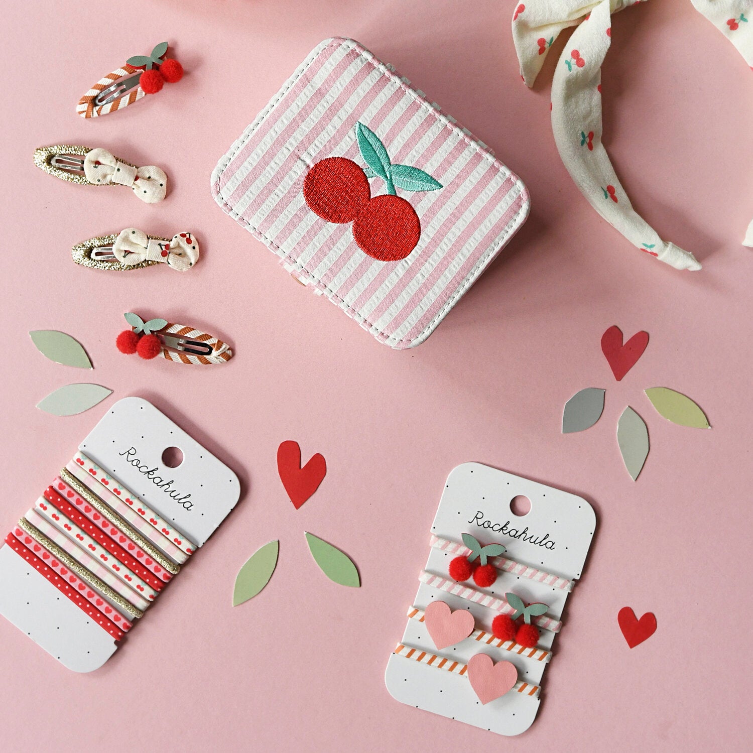 A flat lay on a pink background features the ROCKAHULA - Very Cherry Elastic Pony Pack, along with other Rockahula hair accessories and scattered paper hearts and leaves.