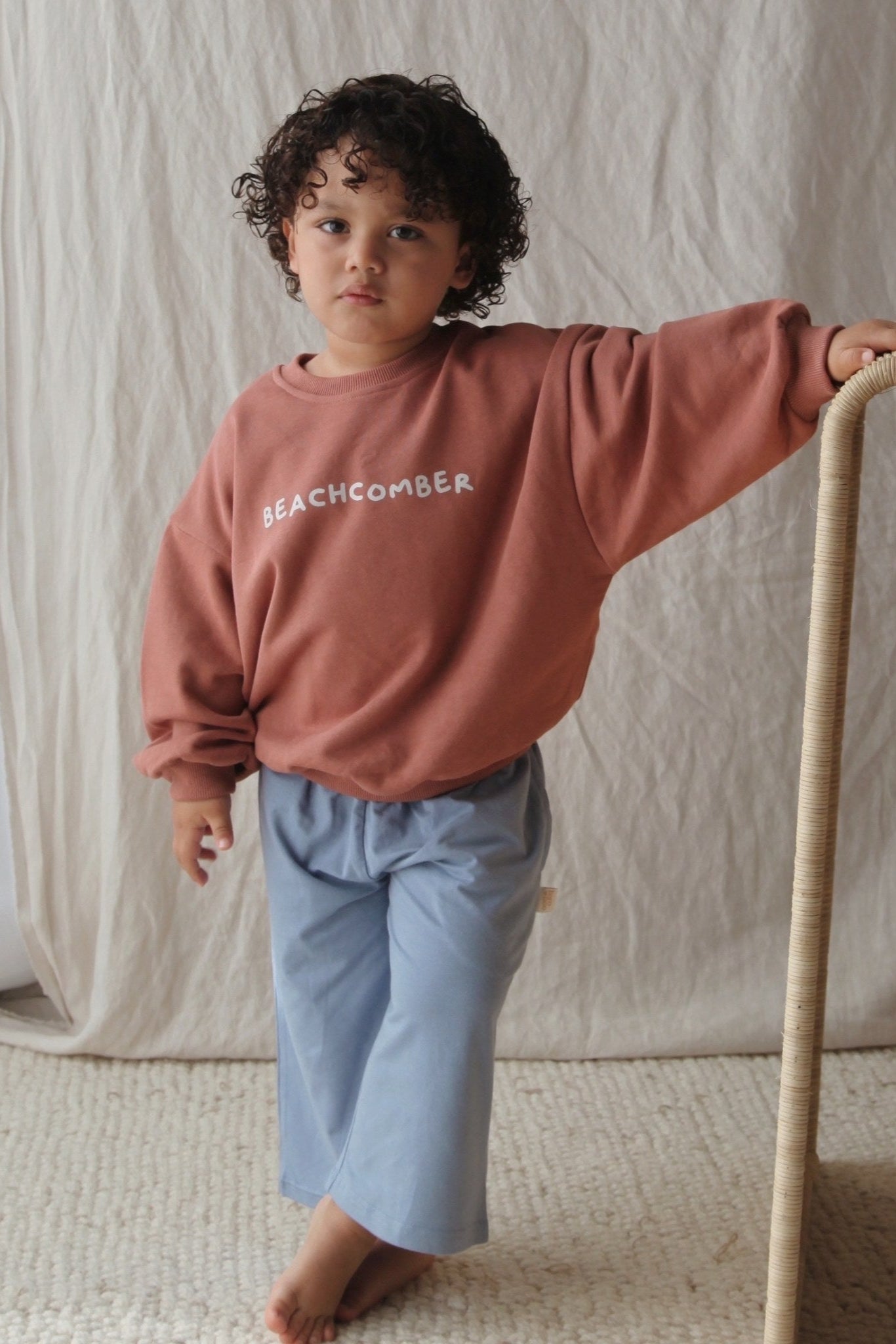 A young child with curly hair stands barefoot indoors, wearing the BAM LOVES BOO Beachcomber Jumper Clay and light blue pants, leaning casually against a woven chair with a neutral fabric backdrop.