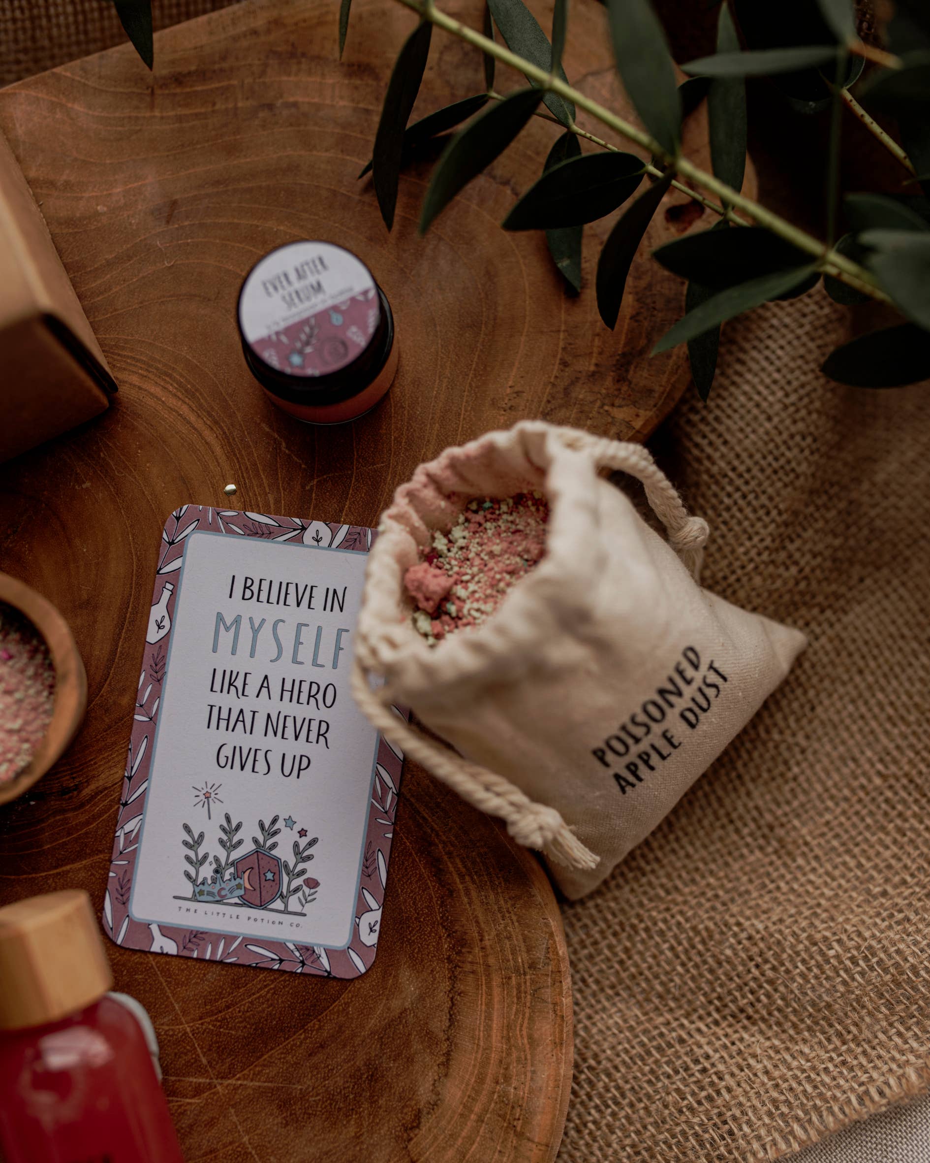 A wooden surface displays The Little Potion Co's MINI Once Upon A Potion Kit, featuring a cloth bag labeled "Poisoned Apple Dust," a card saying “I believe in myself like a hero that never gives up,” and a floral jar—ideal for imaginative play and hinting at enchanted realms beyond the greenery above.