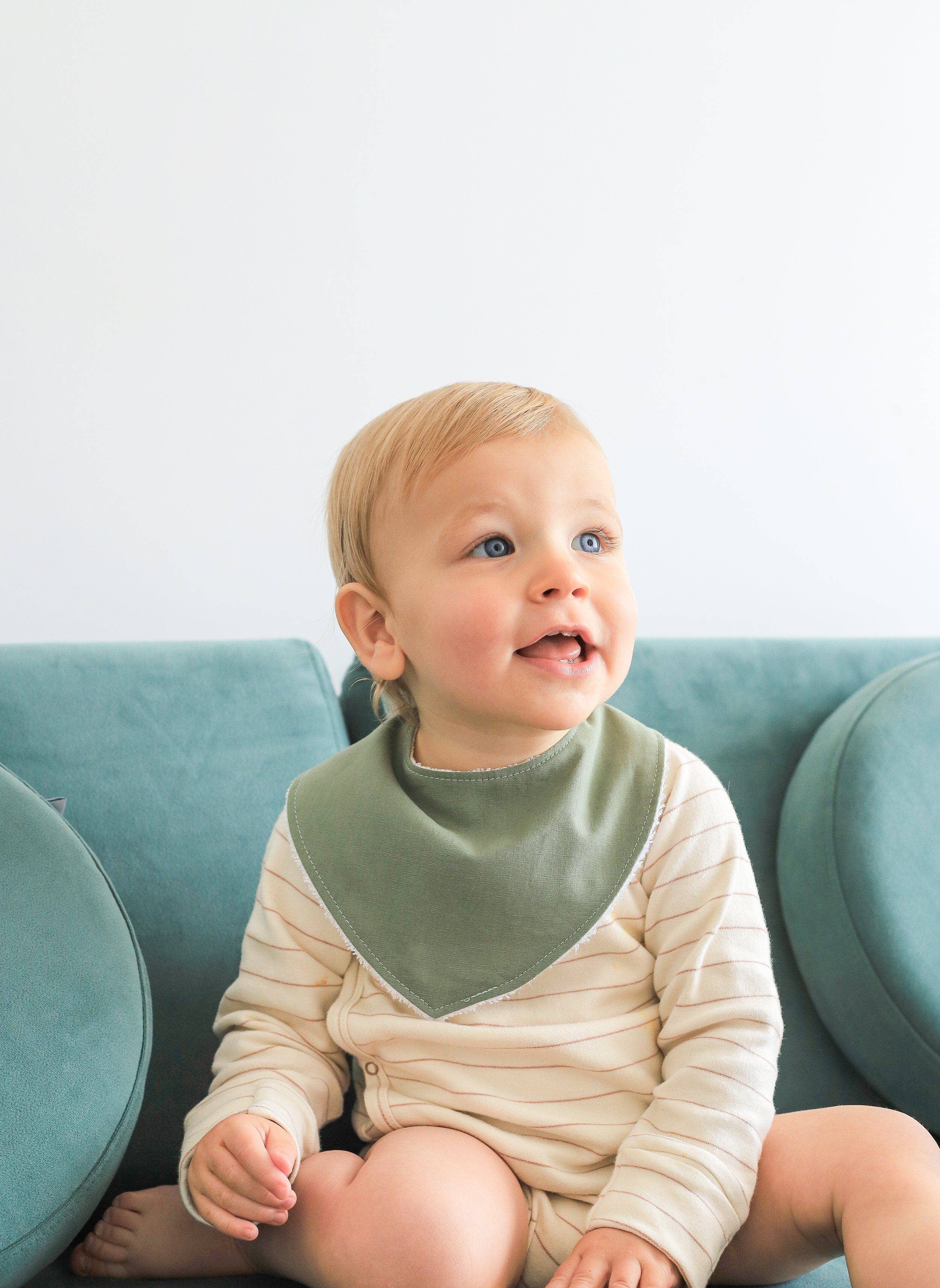 A smiling toddler with light brown hair sits on a teal sofa, wearing a striped onesie and an absorbent Dribble Bib by Kiin Baby. The background is a plain, light-colored wall.