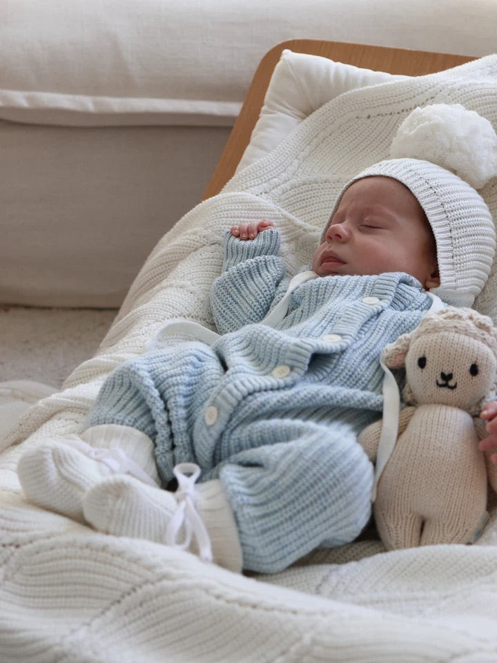 A baby in a ZIGGY LOU Classic Knit Romper Aero Fleck and a white pom-pom knit hat sleeps peacefully on a cream blanket, cuddling a beige stuffed lamb.
