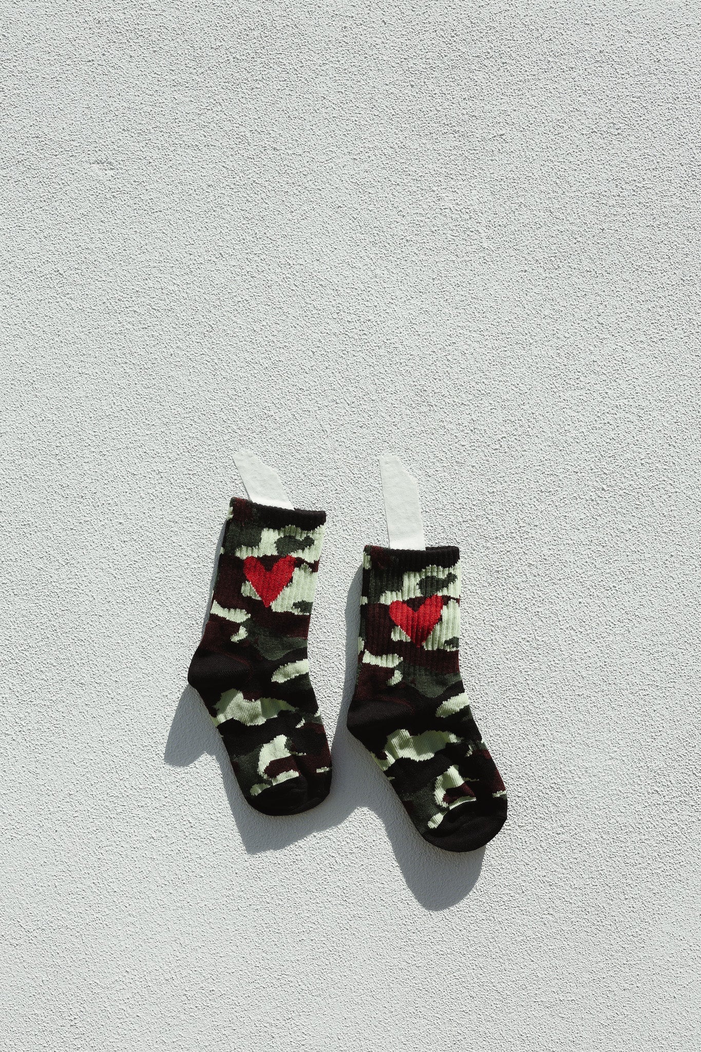TINY LOVE CLUB's Camo Heart Sock for toddlers, featuring a camo pattern with red heart designs and elastic banded ankles, is displayed on a textured white wall, casting soft shadows.