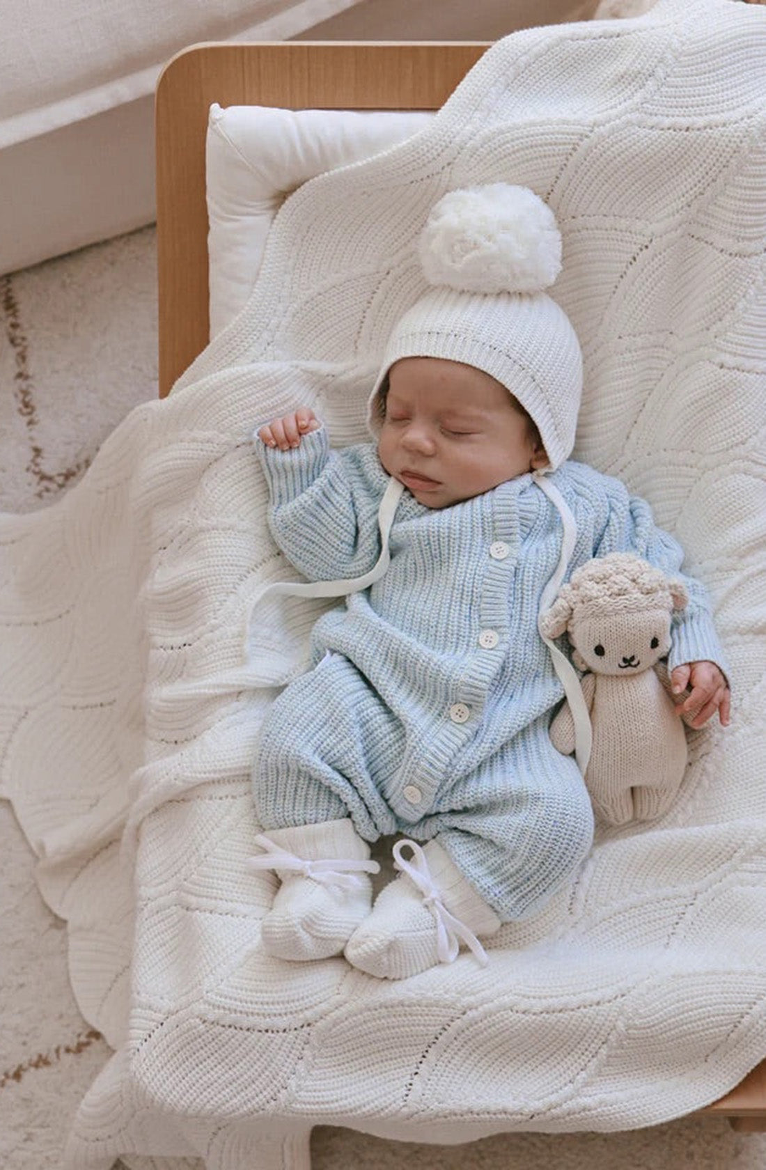 A sleeping baby in a Ziggy Lou - Classic Knit Romper Aero Fleck by ZIGGY LOU and a white pom-pom hat lies on a quilted blanket, cuddling a stuffed animal.
