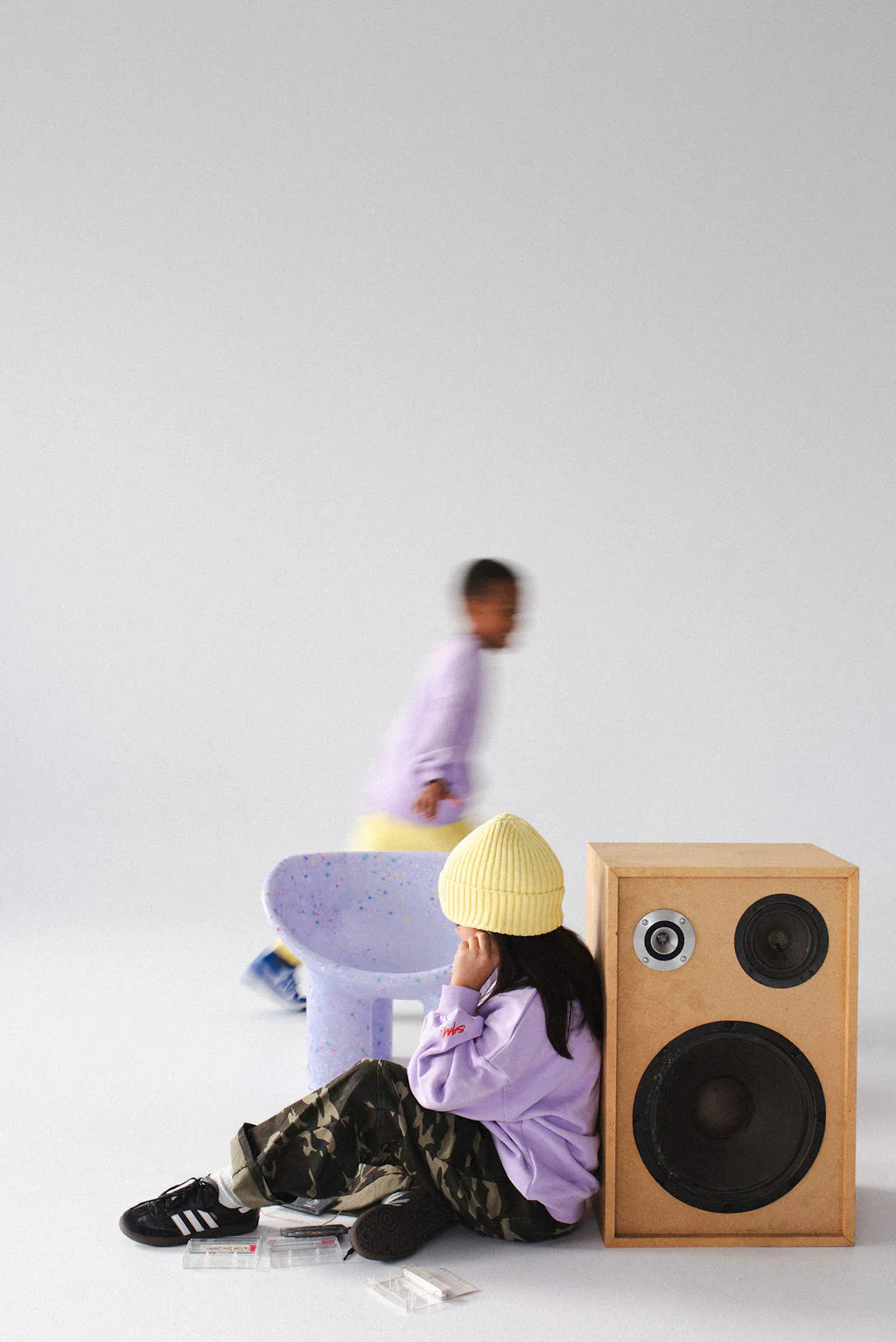 A child in a yellow beanie and Sam the Label - The Cargo Pant Camo from SAM THE LABEL sits by a large speaker, holding a cassette tape. Another child is blurred in motion against a bright, minimal background near a small chair.