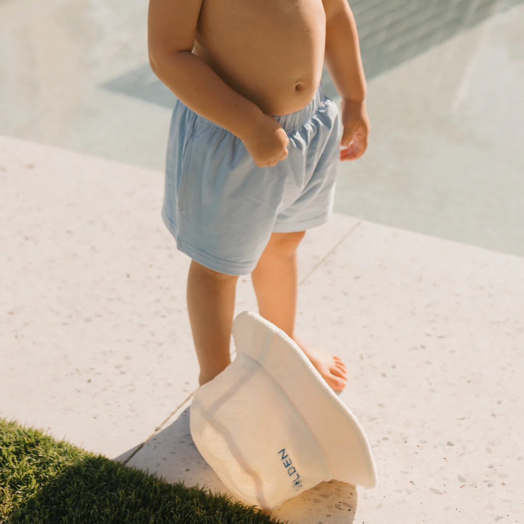 A toddler stands barefoot on light stone by the pool wearing GOLDEN CHILDREN's Retro Boy Shorts Palm Blue. They hold a white sun hat with "EDEN" on it, next to a strip of green grass.