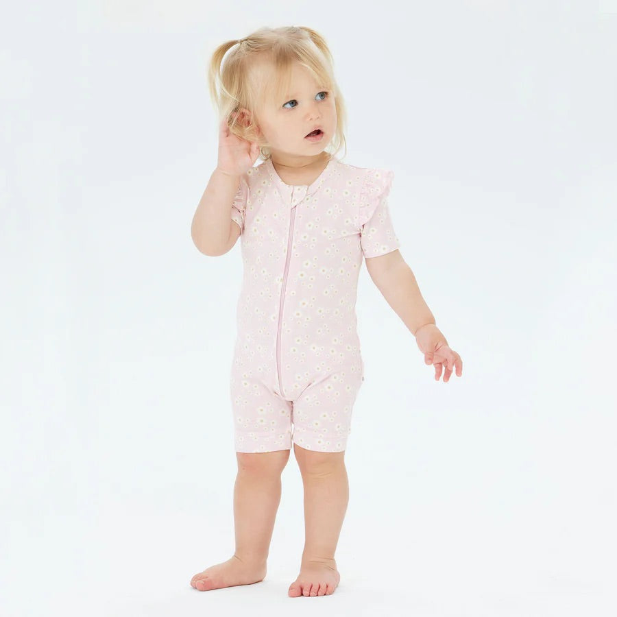 A toddler with blonde pigtails wears the sustainable KYND BABY Day or Night Short Sleeve Frill Onesie in Orchid Bloom, featuring small white flower patterns. The child looks to the side, one hand near the ear and the other arm slightly extended, against a plain white background.