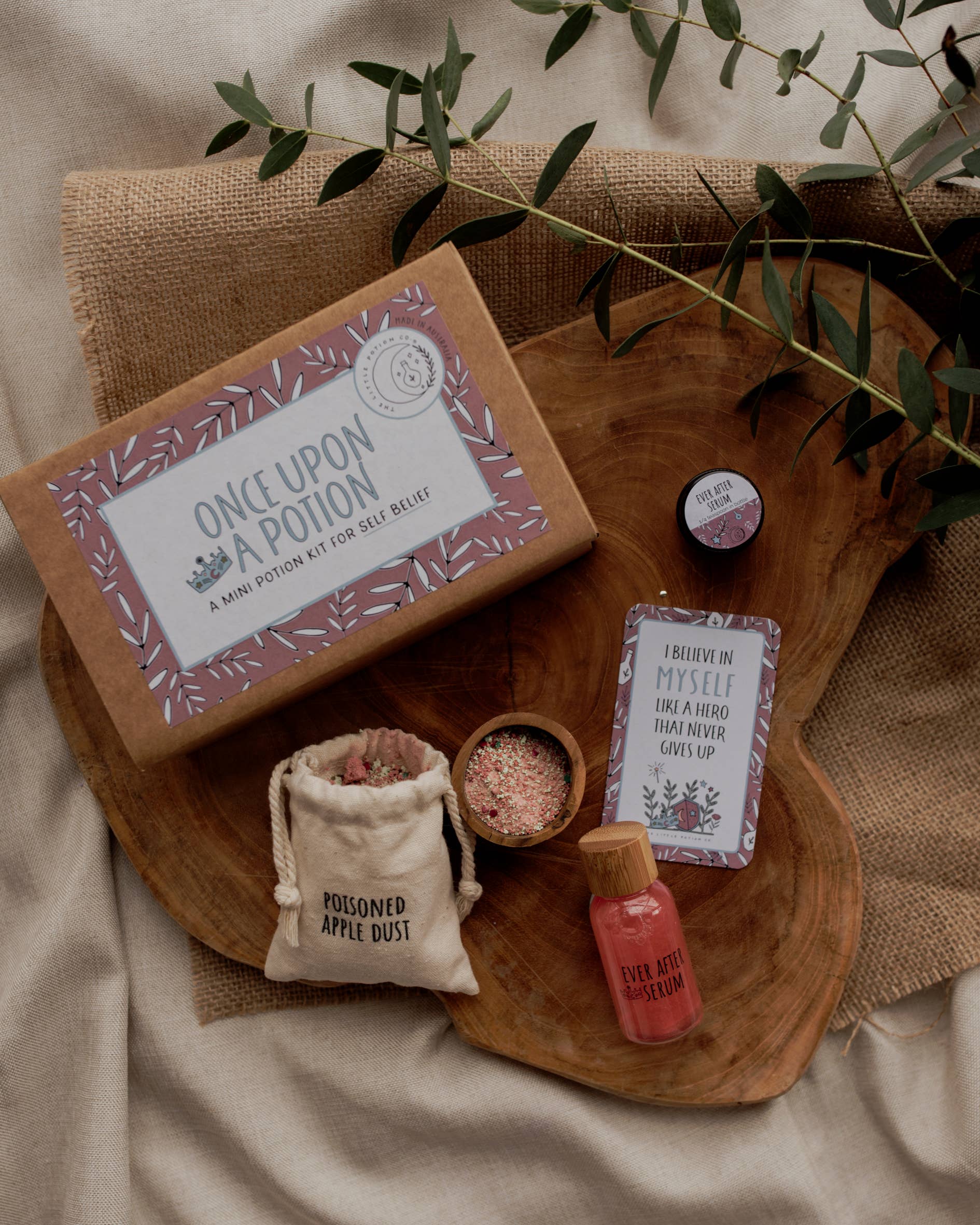 A wooden surface holds The Little Potion Co's MINI Once Upon A Potion Kit, a play set for enchanted realms. Nearby are a small jar, an "Ever After Elixir" bottle, a "Poisoned Apple Dust" sack, an "I believe in myself" card, and scattered greenery.