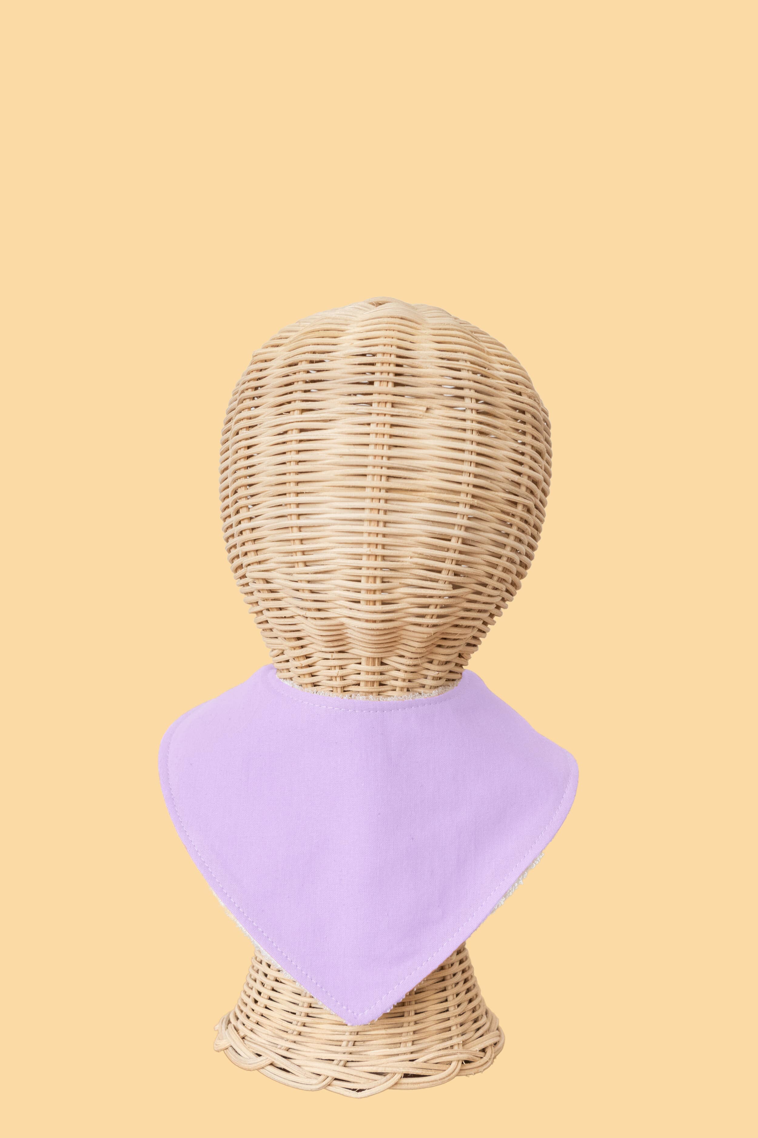 On a light peach background, a wicker mannequin head displays the Kiin Baby Dribble Bib in lavender, made from an absorbent bamboo/cotton blend.