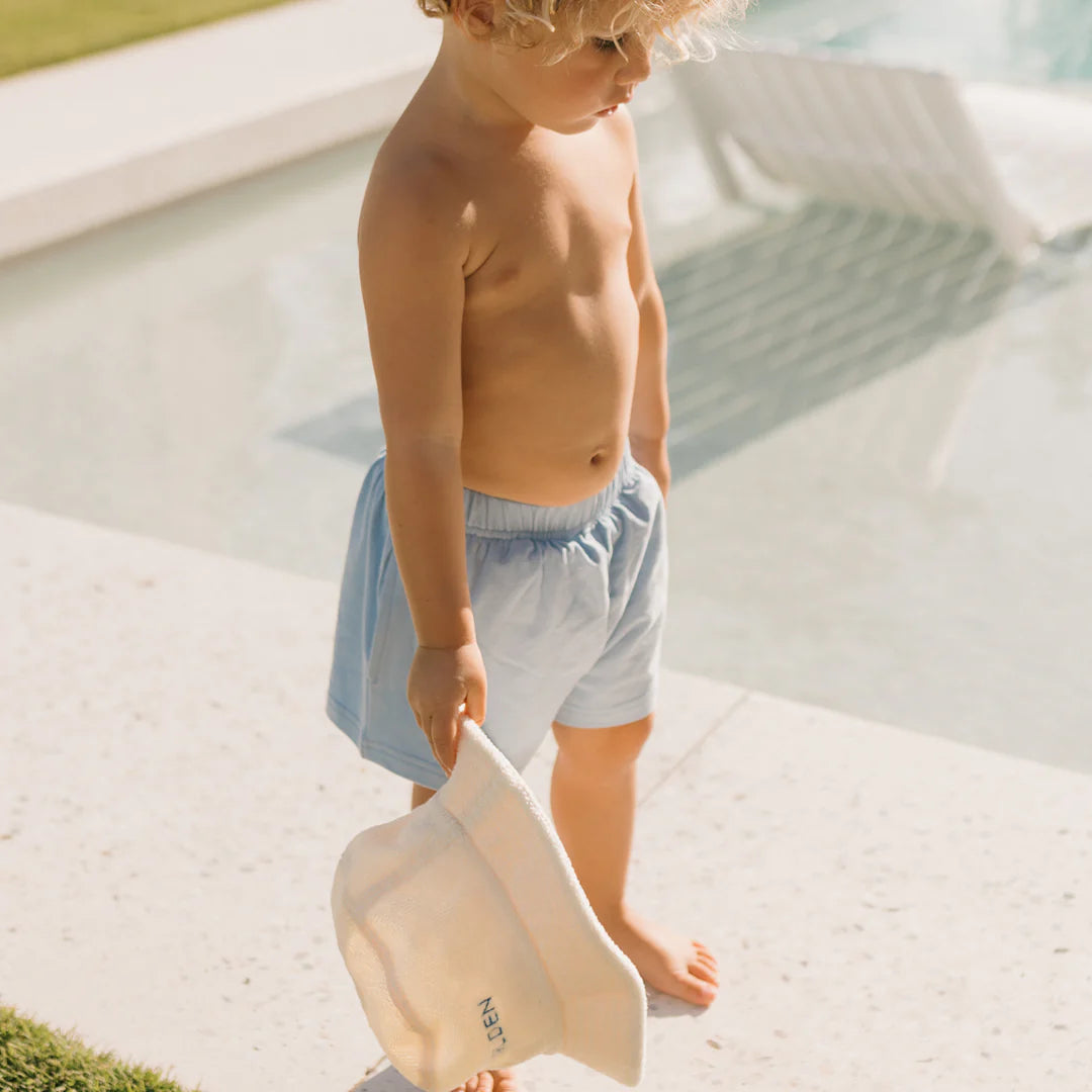 A young child with curly blond hair stands by a pool, wearing GOLDEN CHILDREN's Retro Boy Shorts Palm Blue and holding a white hat. The barefoot adventurer eyes a lounge chair partially submerged in the water as the sun casts its soft light.