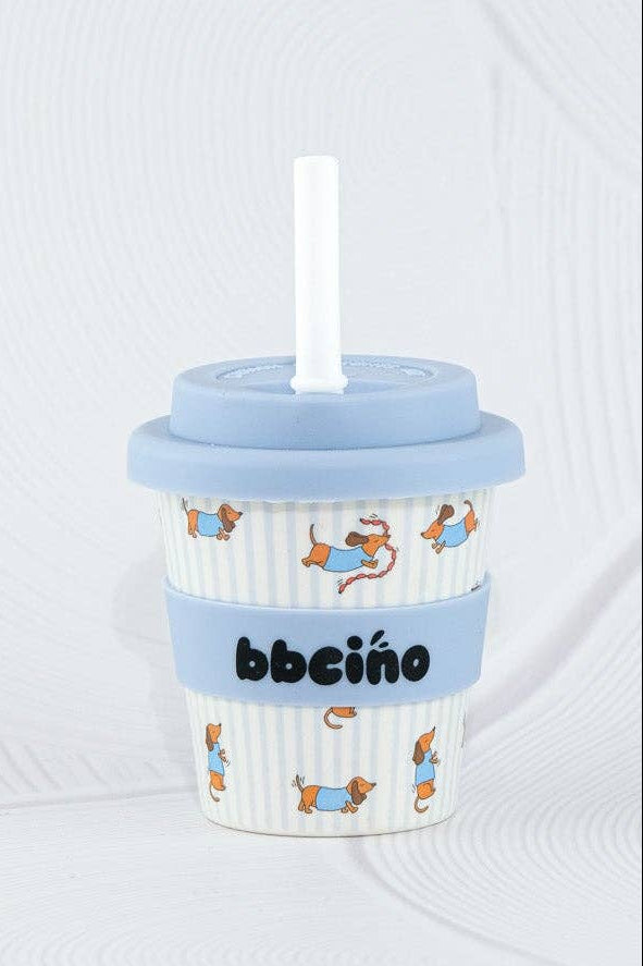 The BBcino - Reusable Babycino Cup 120ml Dash by BBcino features a blue lid, white straw, ribbed blue sleeve labeled “bebeino,” and cute animal designs—an eco-friendly choice for bamboo cup fans. Set against a textured white background.
