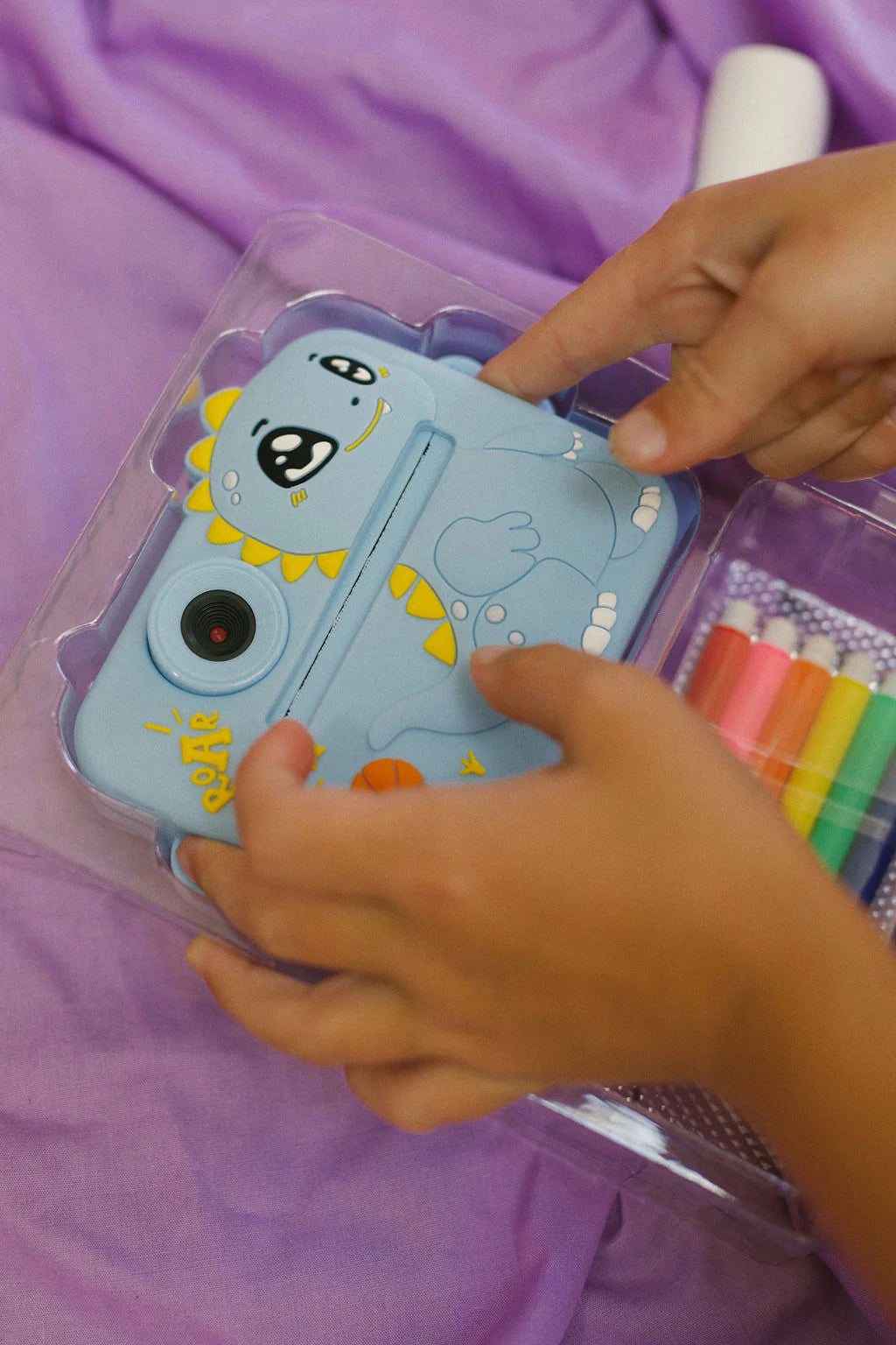 A child's hands adjust the Oh My Frankie - Oh My Snap Instant Print Camera Blue, featuring a cute dinosaur motif. It rests in a clear plastic case on a purple fabric background beside colorful markers, sparking creativity for kids.