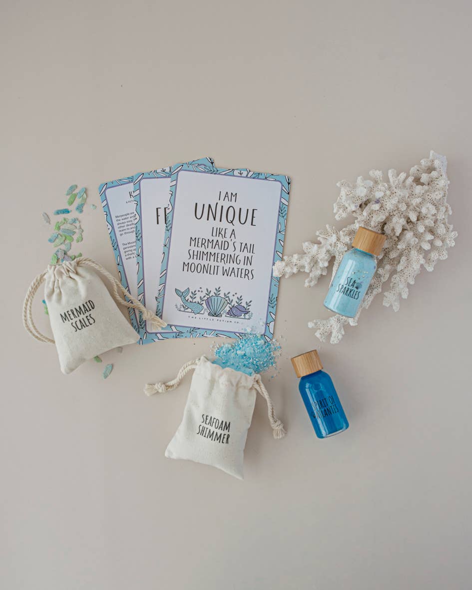 A flat lay image featuring three inspirational quote cards, the Moonlight Waters MINDFUL Potion Kit by THE LITTLE POTION CO with two small drawstring bags labeled "Mermaid Scales" and "Seafoam Shimmer," two small bottles of blue liquids, iridescent confetti, and a piece of white coral. The theme is mermaids and ocean beauty.