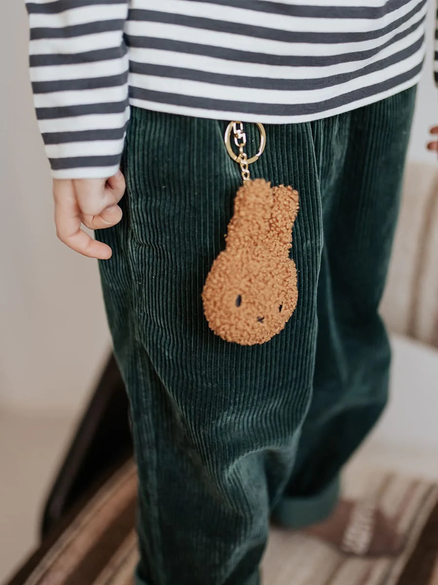 A child wearing green corduroy pants and a black-and-white striped shirt has a small, brown **BON TON TOYS X MIFFY** Miffy Flat Keychain Tiny Teddy Cinnamon made from recycled polyester attached to their pants near their upper thigh.