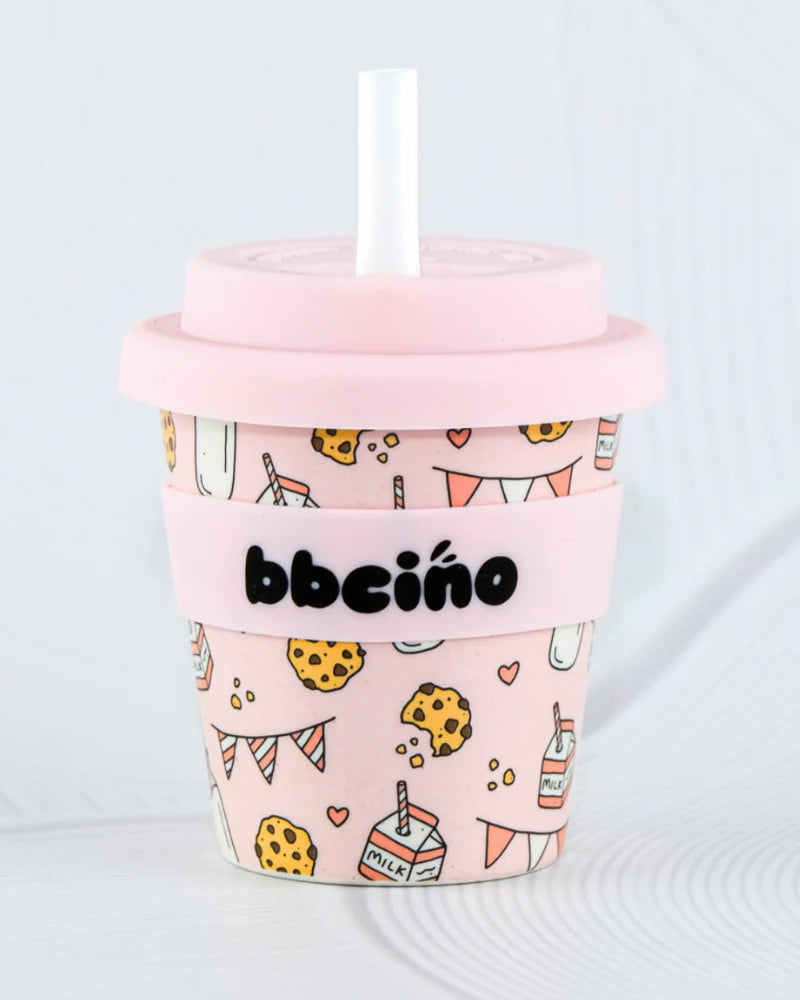 The BBcino Reusable Babycino Cup 120ml in Tough Cookie Pink features a pink lid, white straw, and sleeve labeled "bbcino." Decorated with cookies, milk cartons, and hearts, it's made from sustainably farmed bamboo for an eco-friendly touch.