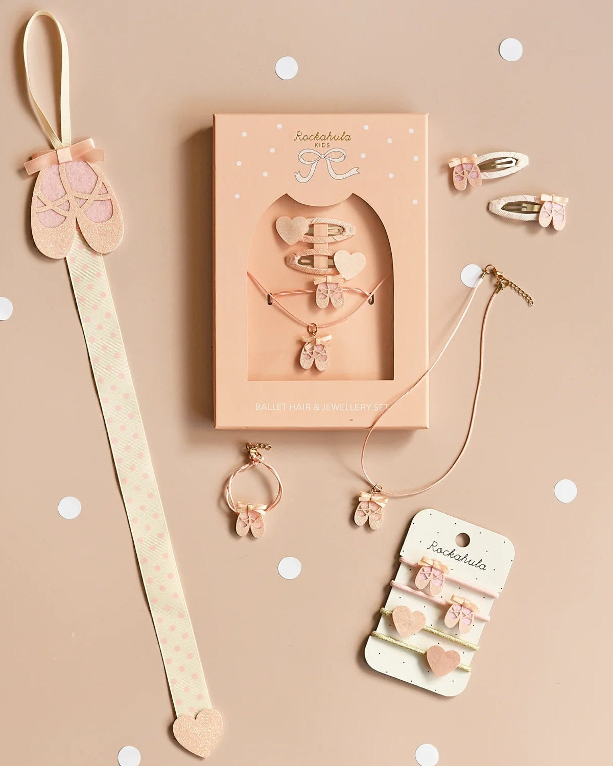 A flat lay of ROCKAHULA’s Rockahula - Ballet Shoes Ponies, pink ballet-themed accessories arranged on a beige surface with white dots, including hair ties decorated with glittery pink ballet slippers.