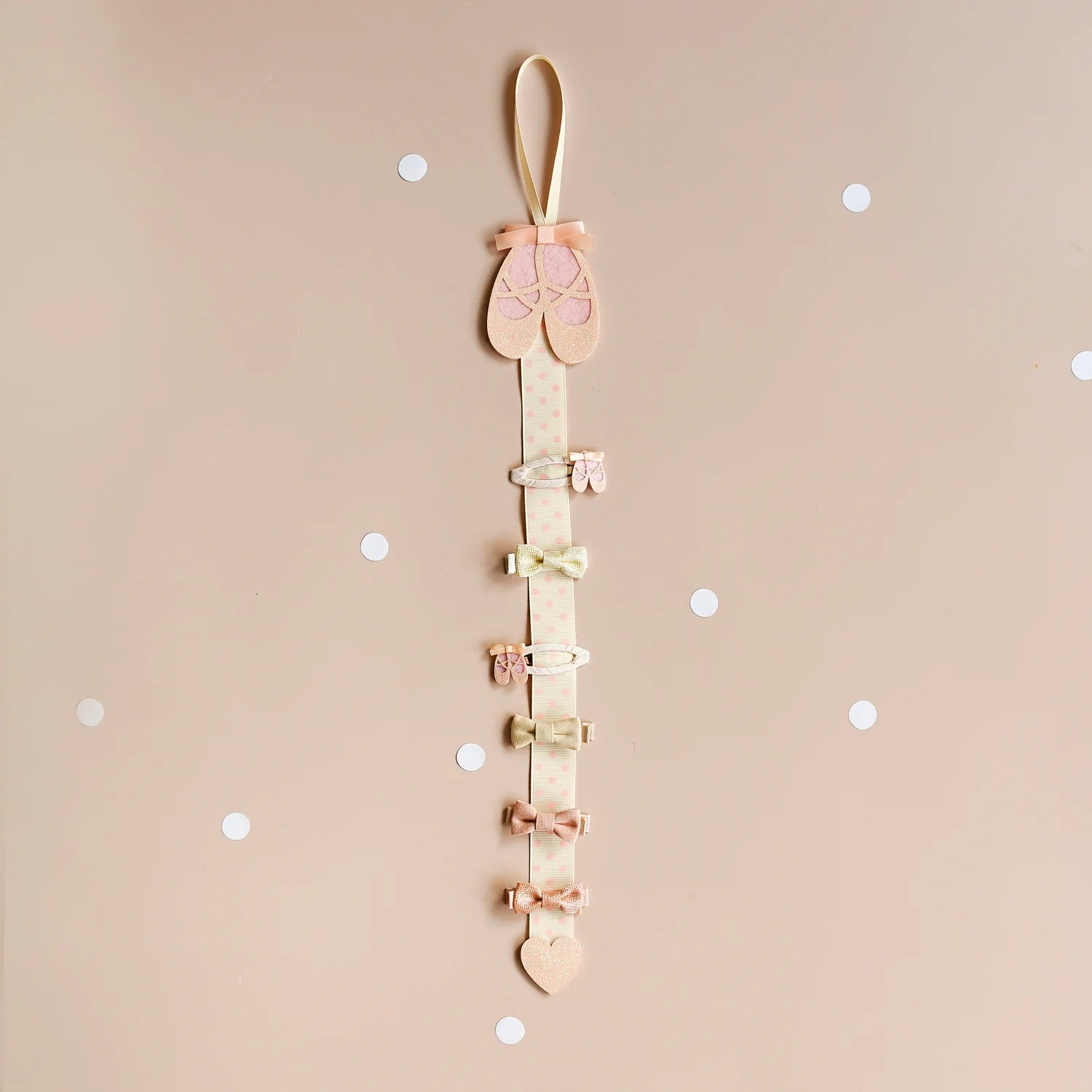 The ROCKAHULA Rockahula - Ballet Shoes Clips is a vertical hair clip organizer topped with pink glitter ballet slippers, holding six pastel bow snap clips on a ribbon, shown on a beige background with white dots.