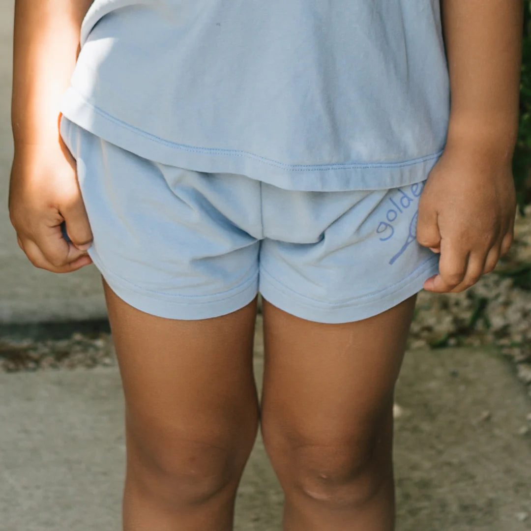 A child stands on a path wearing a light blue cotton jersey shirt and Retro Boy Shorts Palm Blue by GOLDEN CHILDREN. The shorts say "gold" in purple on the lower right side, with an elasticated waist. Greenery and concrete are in the background.