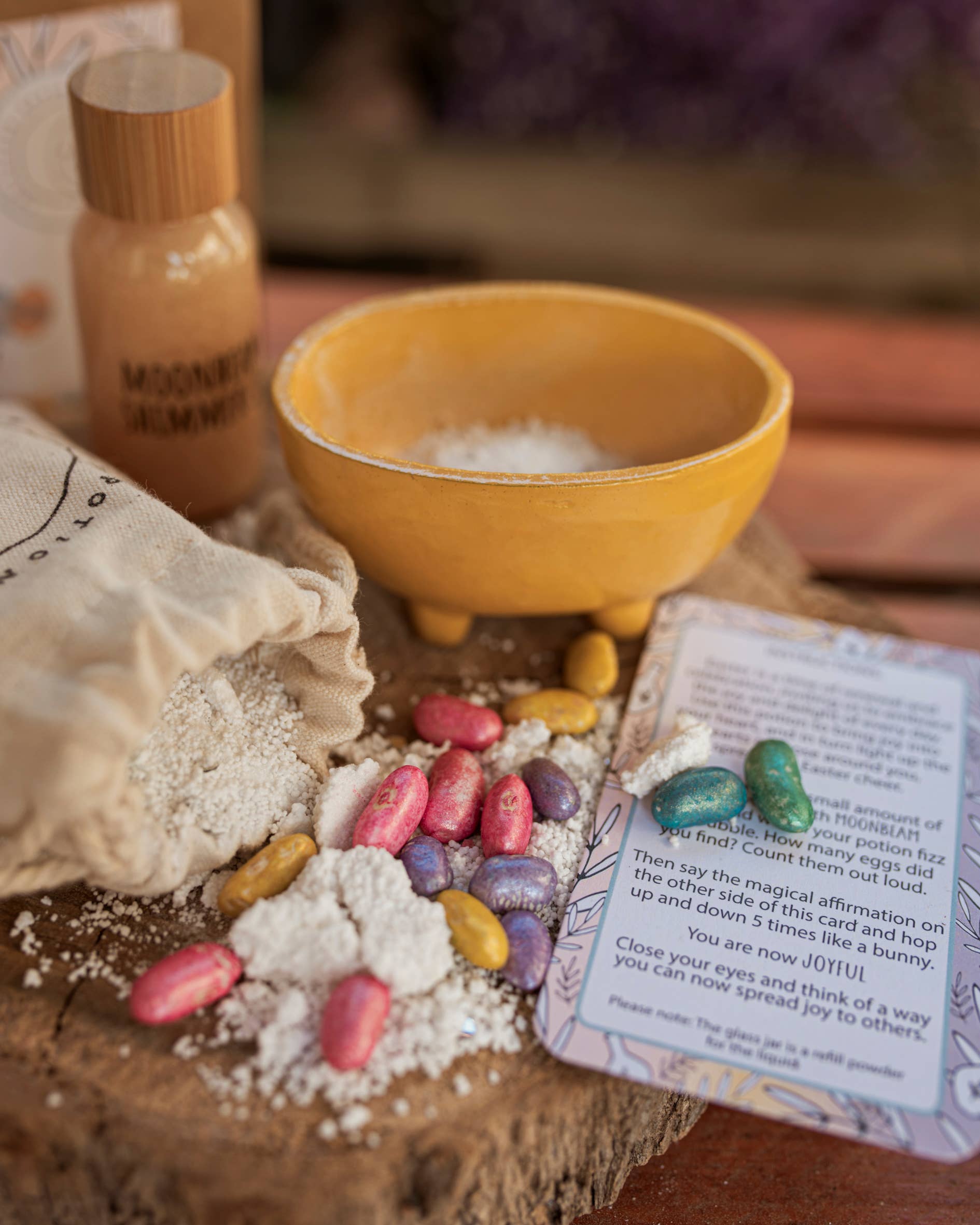 A rustic display presents The Little Potion Co's MINI Easter Egg Hunt Potion Kit, featuring a yellow bowl of powder, colorful oval candies, a text card, a "Moonlight Shimmer" bottle, and a powder-filled cloth bag on wooden surface.