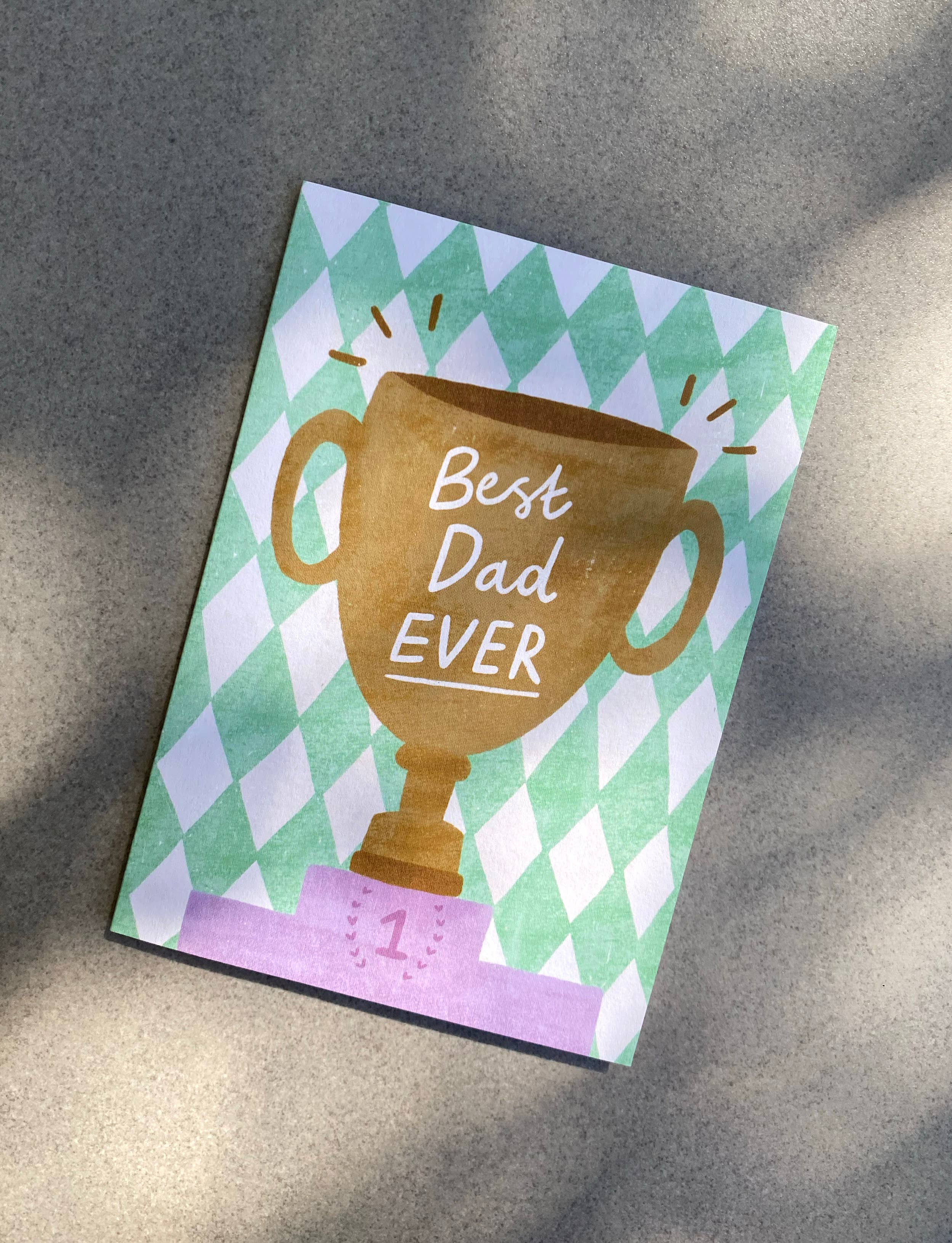 The 'Best Dad Ever' Trophy Father’s Day Greeting Card by LAUREN SISSONS STUDIO features a gold trophy, pink ribbon with "1", and "Best Dad EVER" on a green and white diamond background—an ideal recycled card to celebrate dad. Shown on a gray surface.