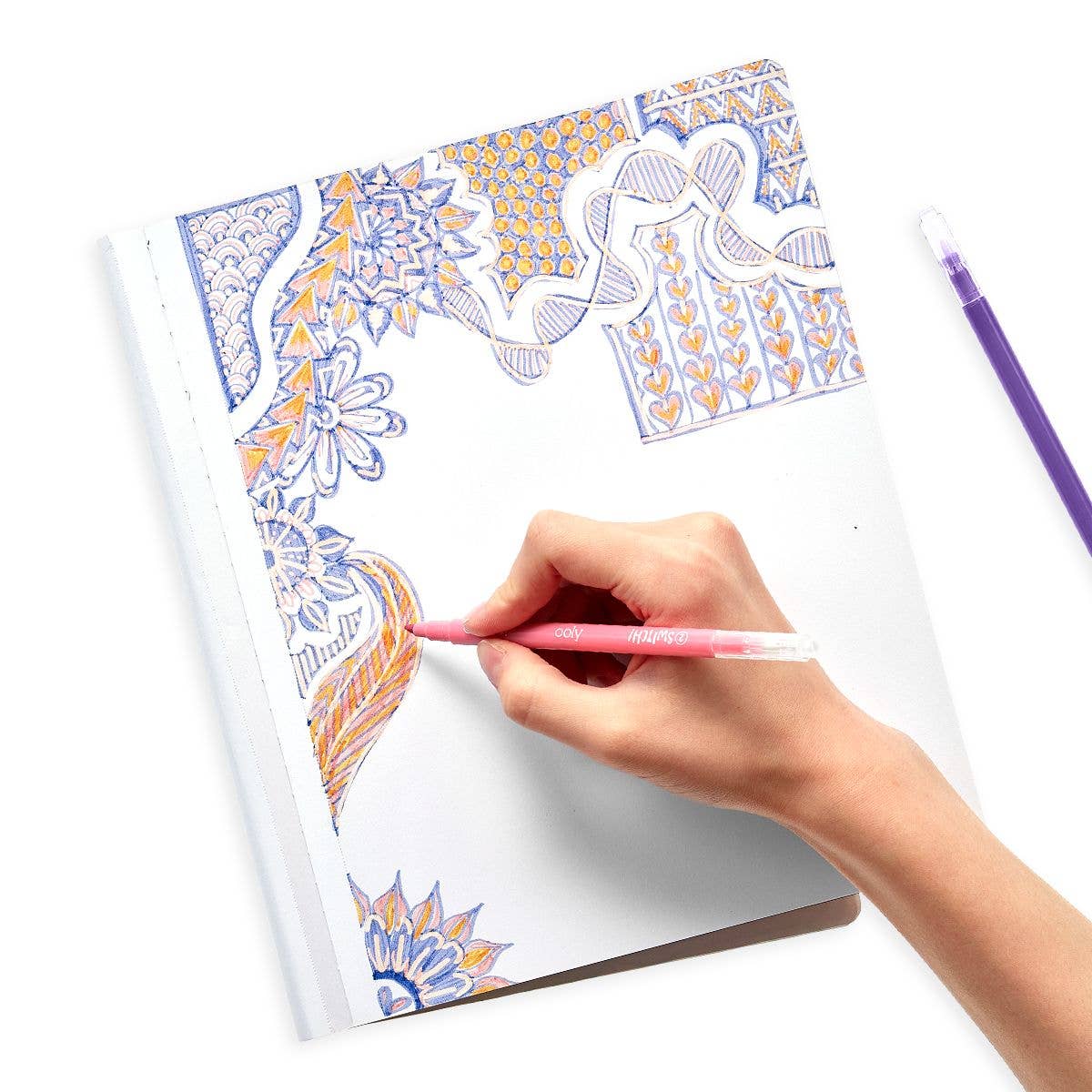 A person draws detailed floral and abstract patterns in orange, blue, and yellow on white paper with a red pen. A purple pen and OOLY's Switch-eroo! Color-Changing Markers 2.0 lie nearby among other art tools, ready for creative inspiration.